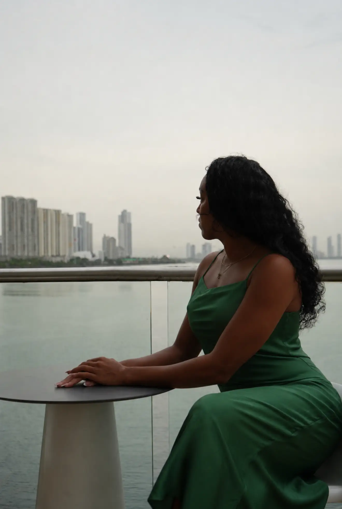 Person in a green dress sits at a balcony table overlooking a waterfront city skyline.