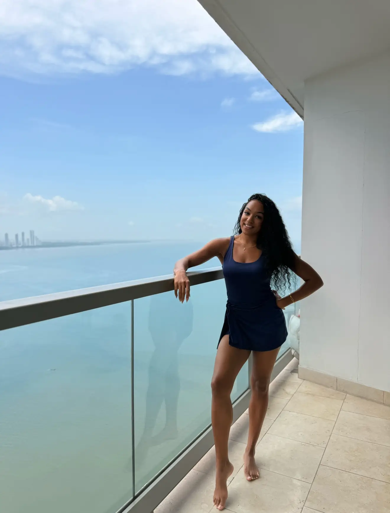 Person in a navy outfit stands barefoot on a high balcony overlooking blue water and a distant skyline.