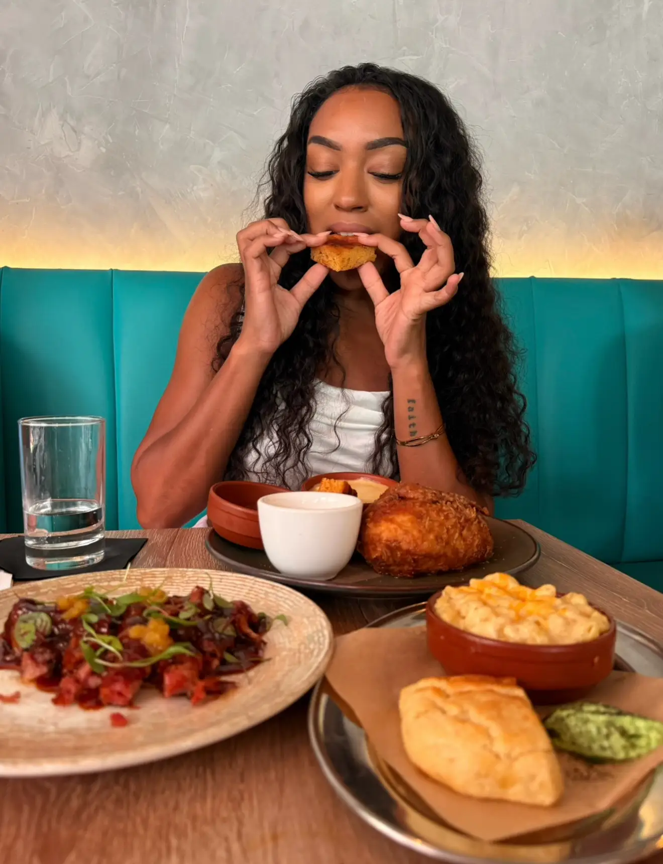 A person seated in a restaurant booth bites into fried food with multiple plates of food on the table.