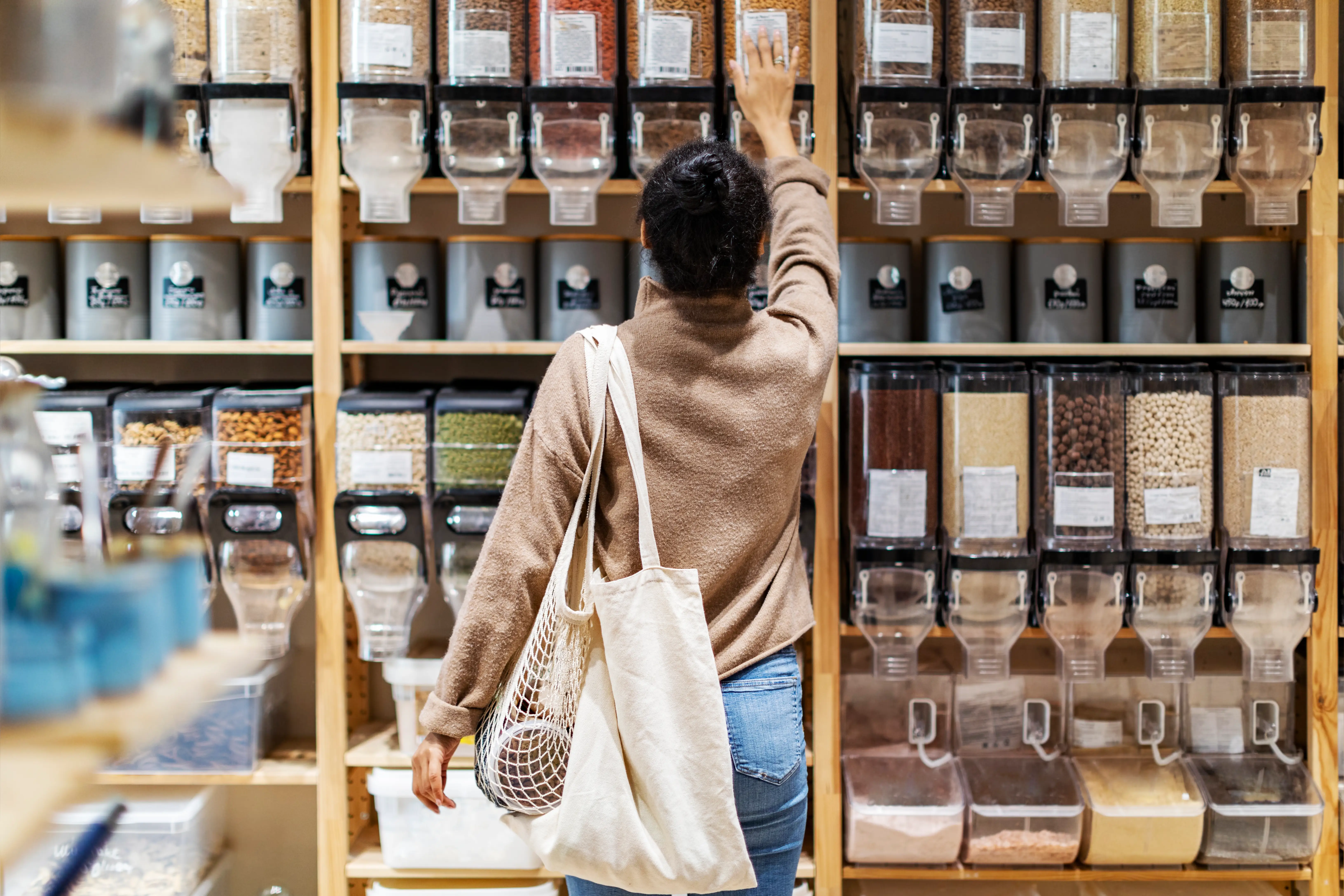 Shopper with a tote bag reaches toward bulk dry goods dispensers in a grocery store aisle.