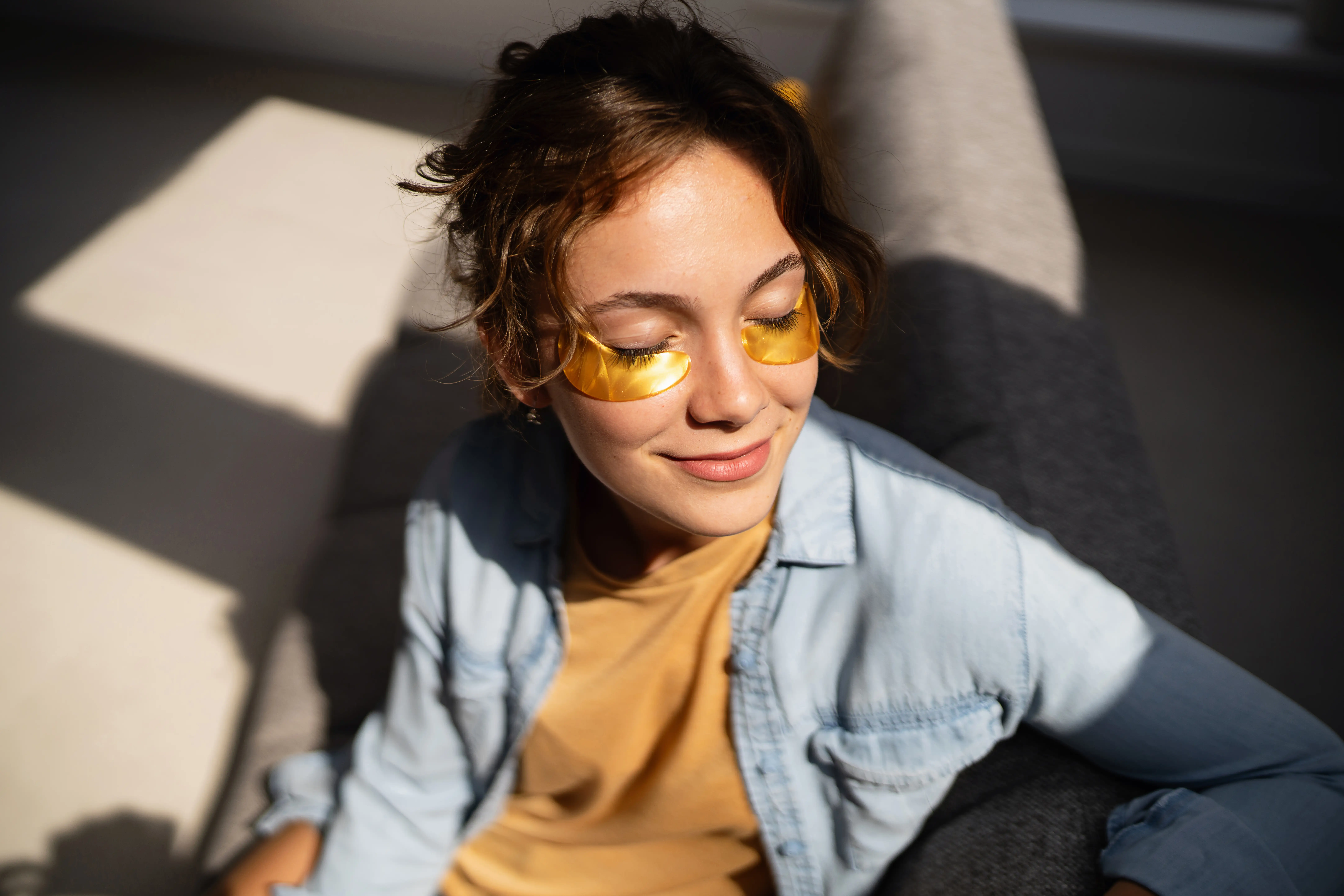 Person relaxes in warm sunlight wearing yellow under-eye patches and a light denim shirt on a gray chair.