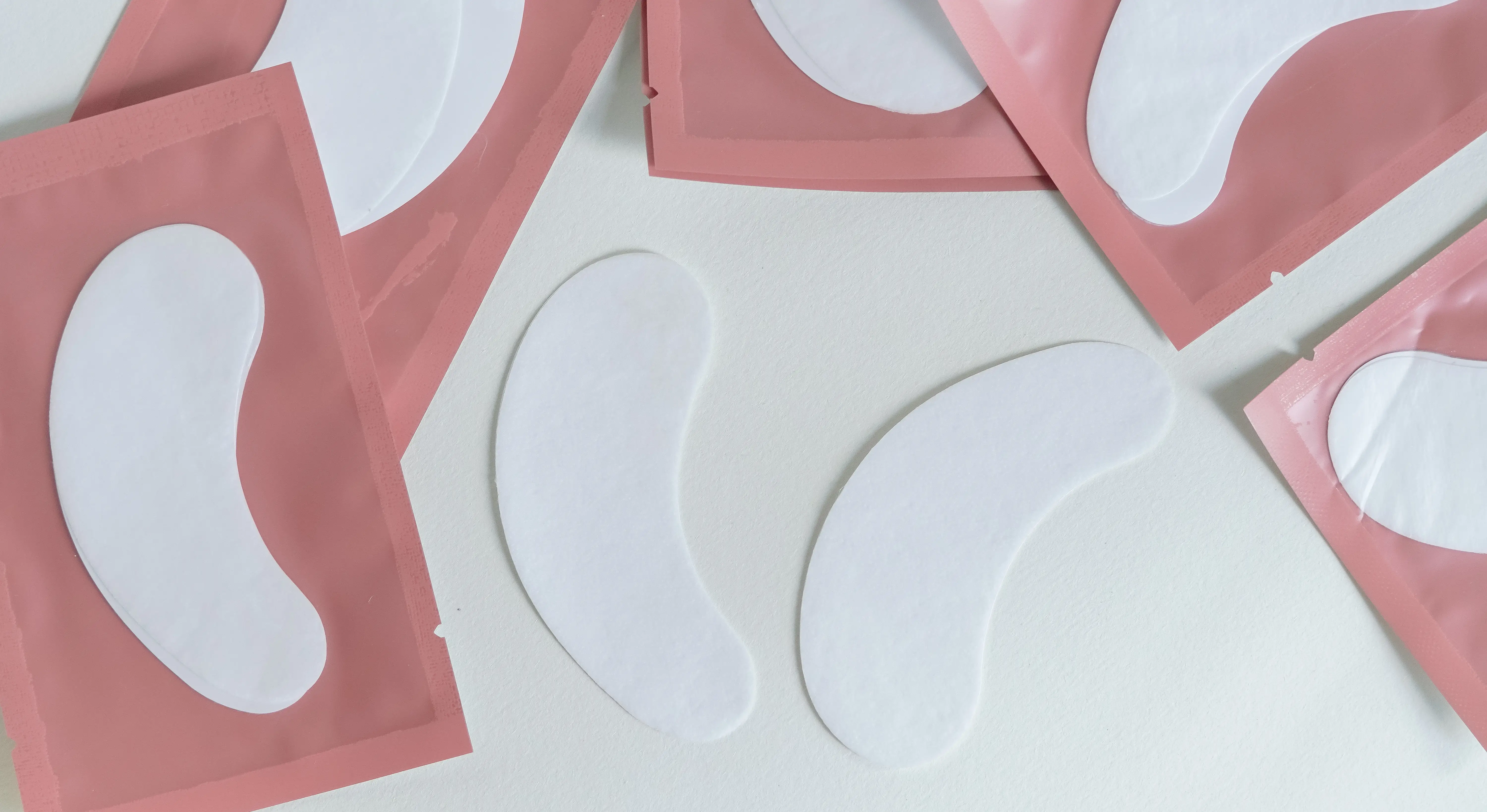 White under-eye gel patches and pink cosmetic sachets are arranged on a pale surface.
