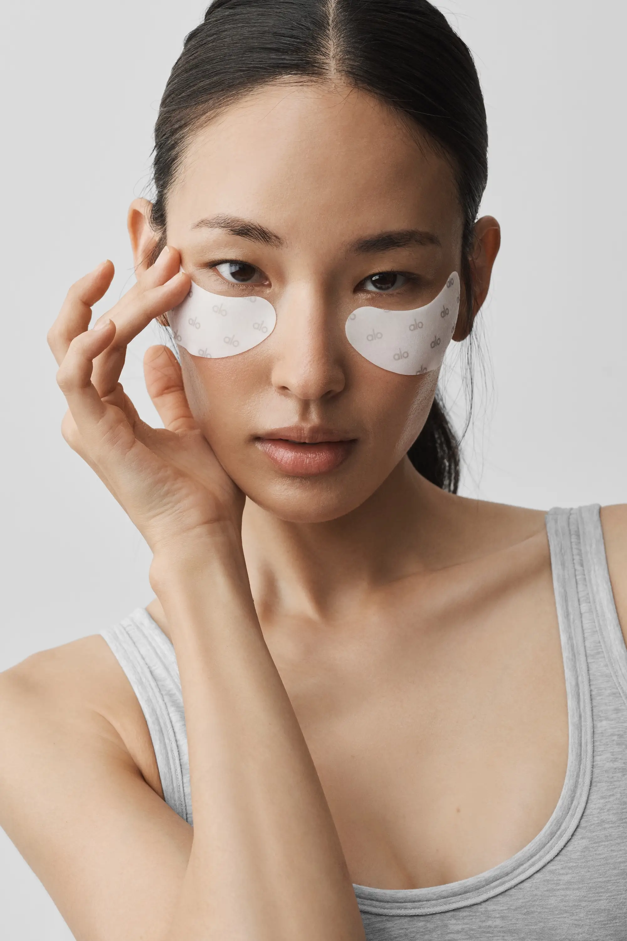Person wearing white alo under-eye patches and a gray tank top touches their face against a plain light background.