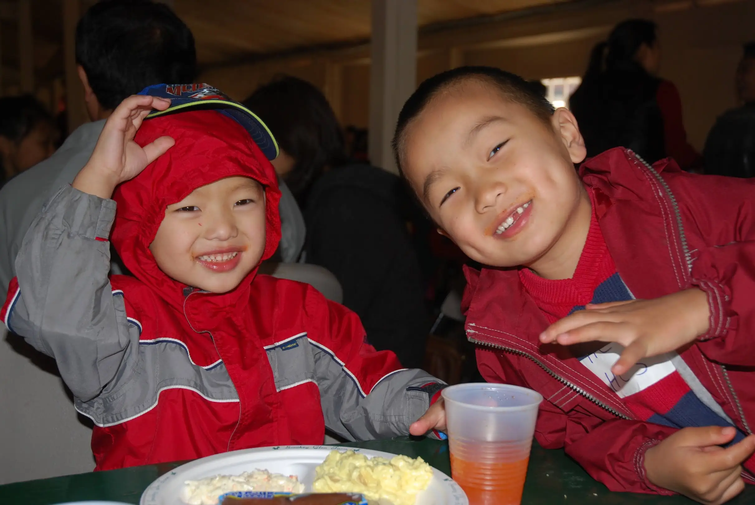 Two smiling people in red jackets sit at a table with food and a plastic cup indoors.