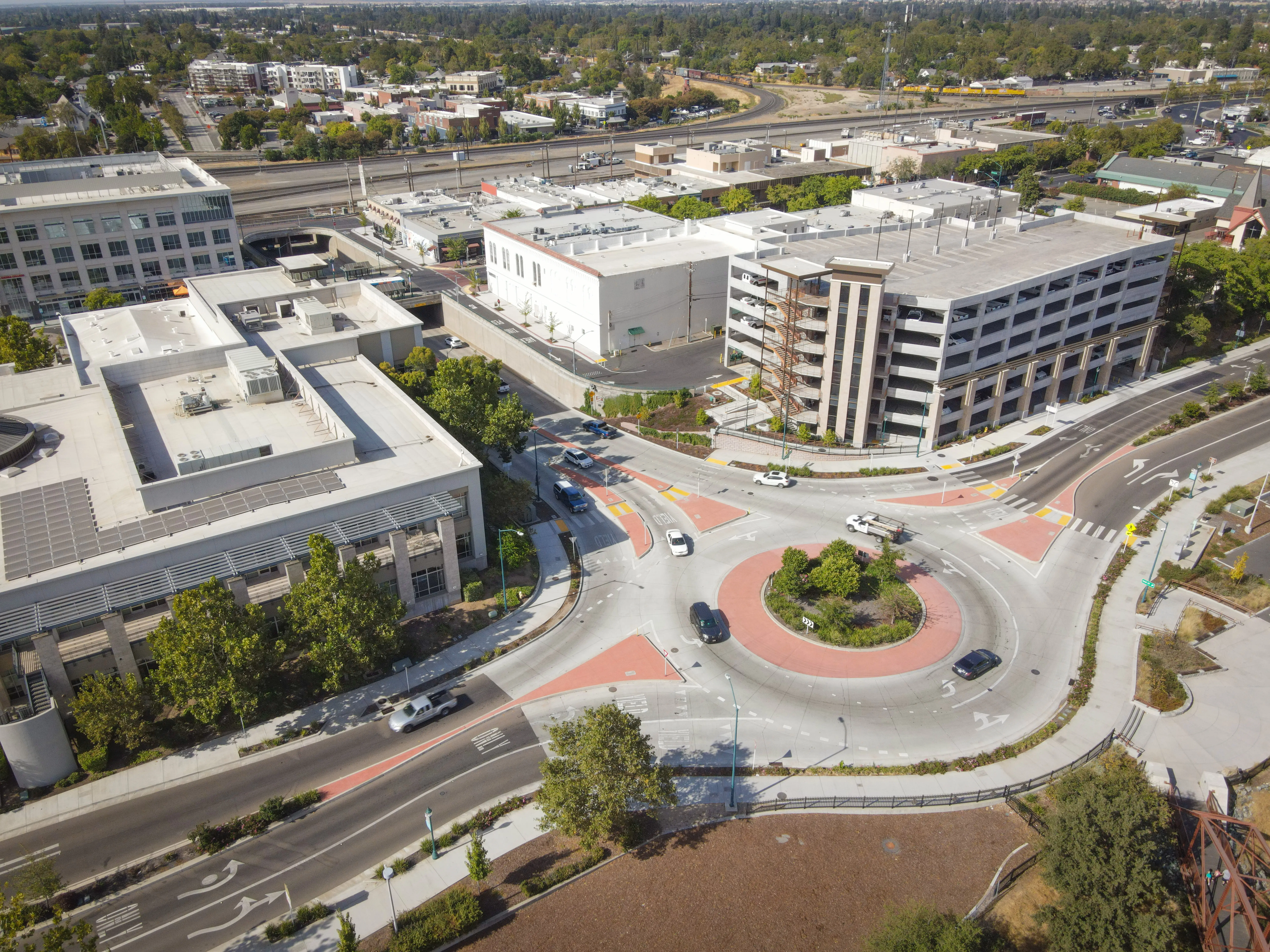 Aerial view of an urban roundabout surrounded by office buildings, streets, parked cars, trees, and rail tracks.