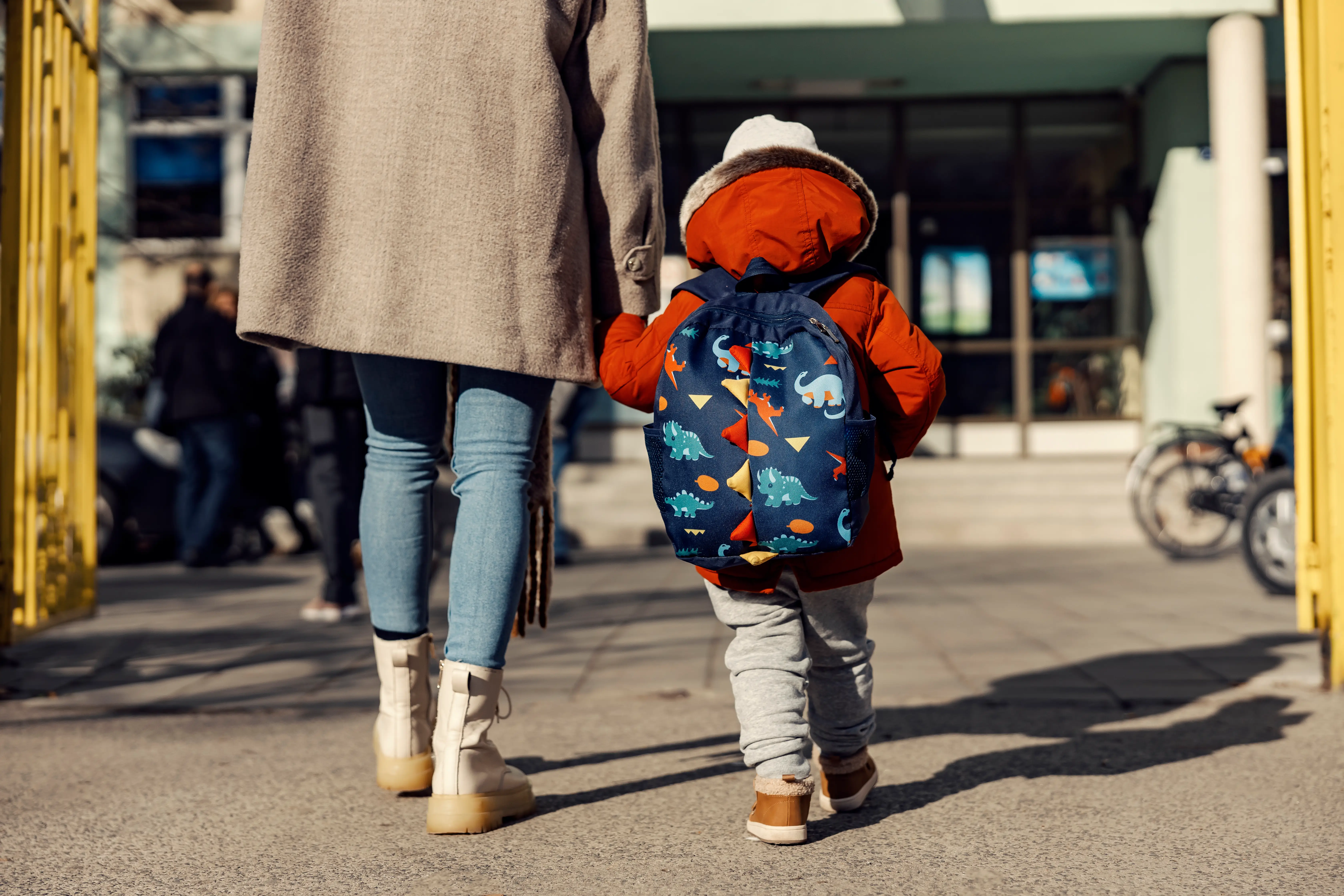 Small child with a colorful backpack walks hand in hand with an adult toward a building entrance.