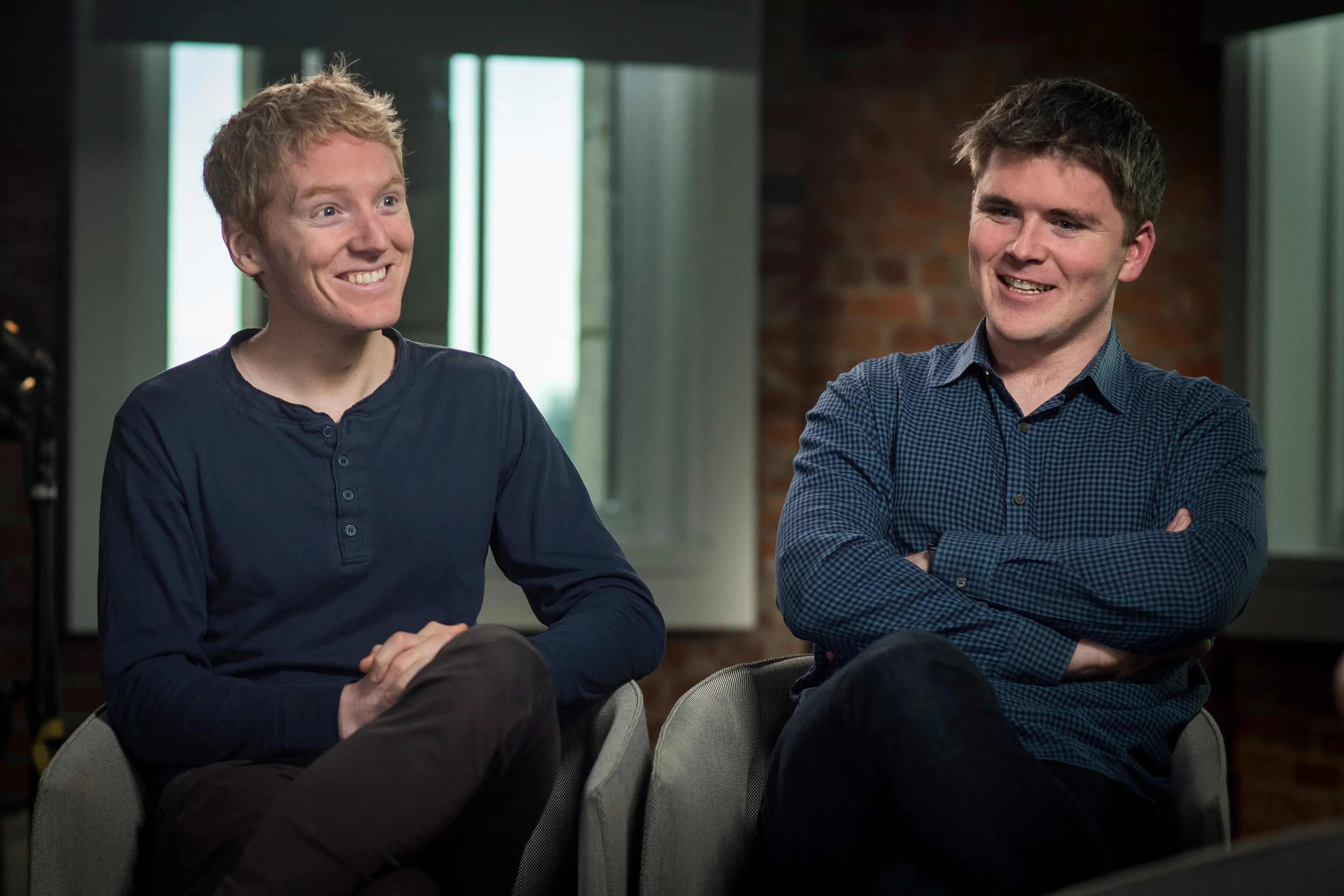 John Collison and Patrick Collison sit in armchairs during an indoor interview in a brick-walled room.