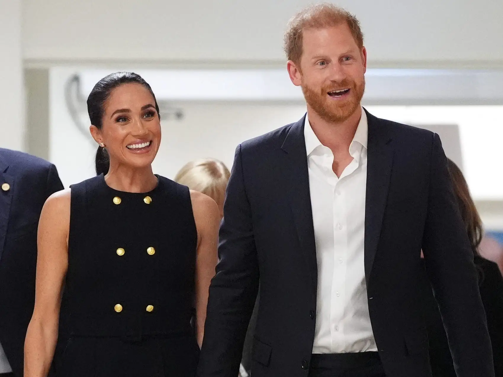 Meghan Markle and Prince Harry walk hand in hand through an indoor corridor in formal dark attire.