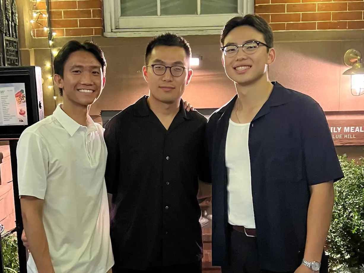 Three people pose together outside a brick-front restaurant entrance decorated with string lights at night.