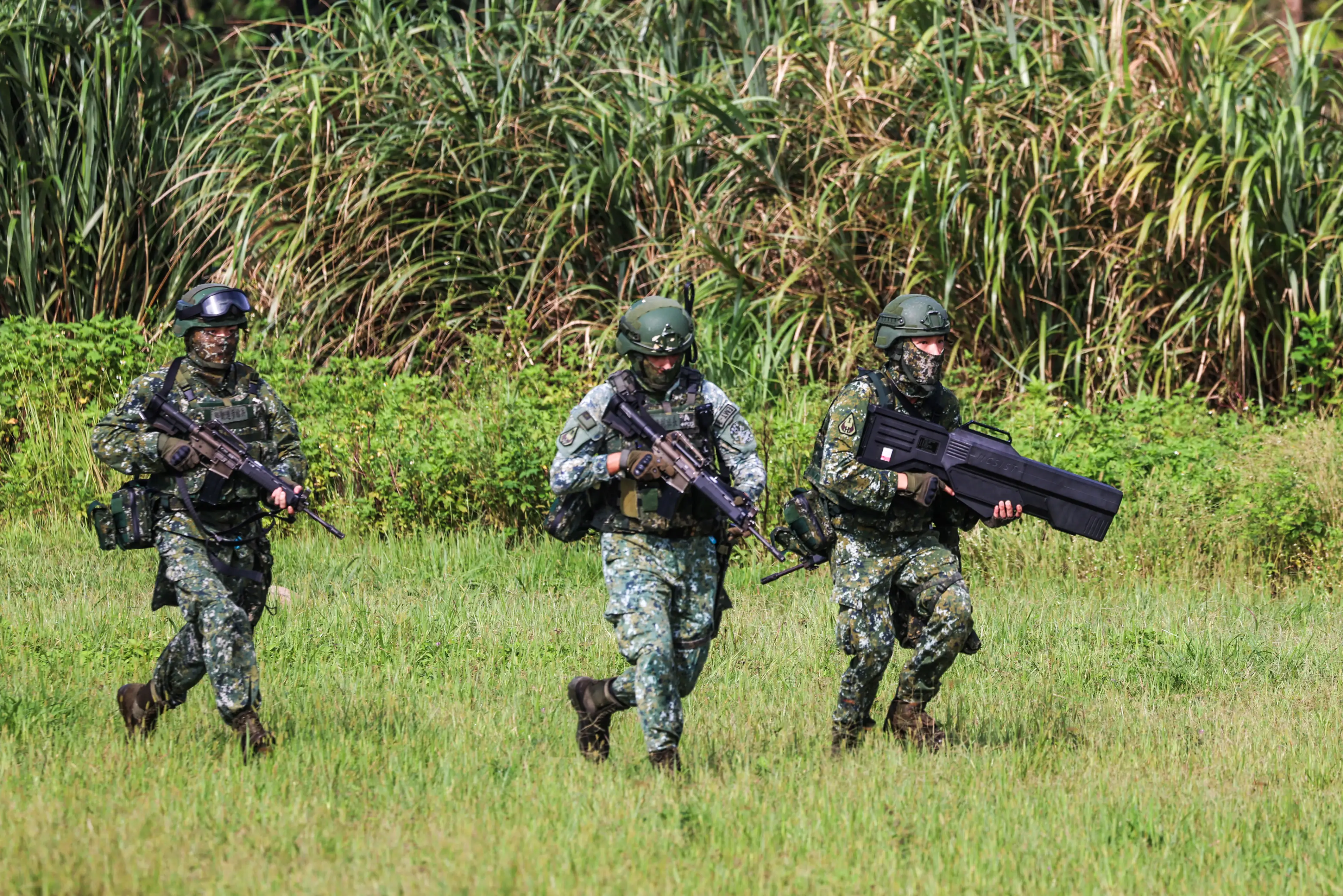 Three Taiwanese soldiers with rifles and an anti-drone gun run on the grass.