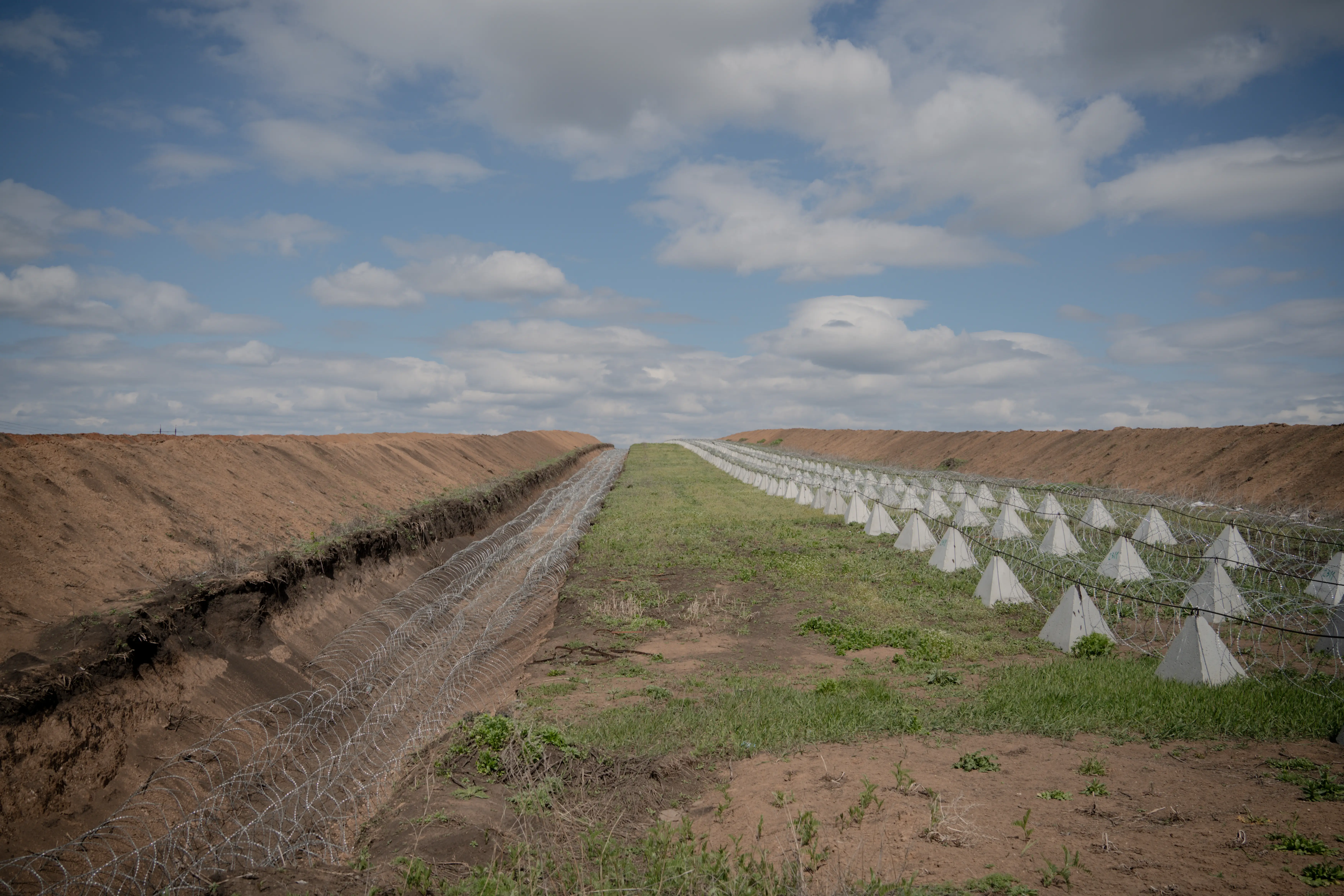 Fortified earthen trench with barbed wire and concrete pyramid barriers stretches across an open landscape under cloudy sky.