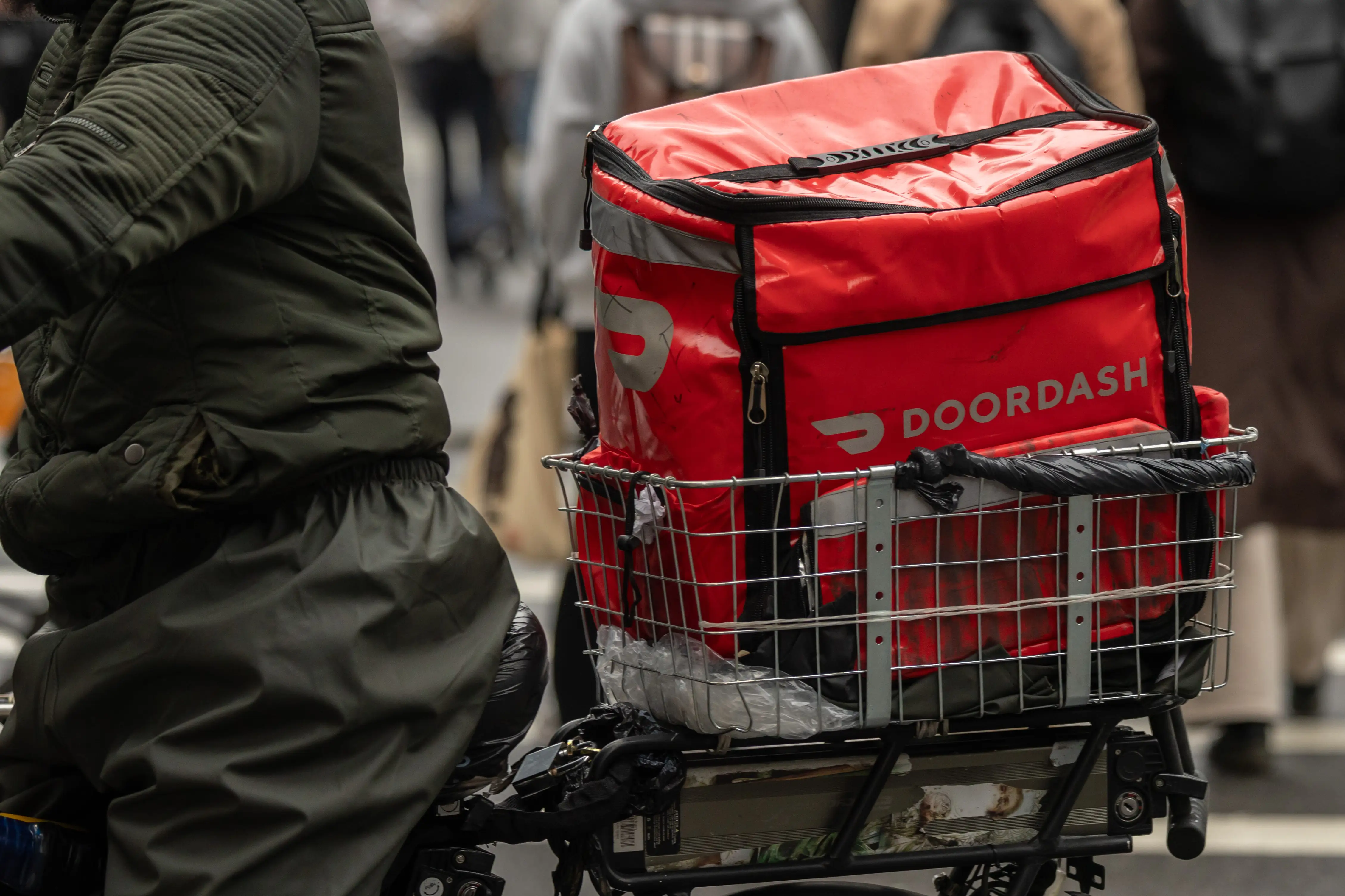 Red DOORDASH insulated delivery bag sits in a rear bicycle basket on a crowded city street.