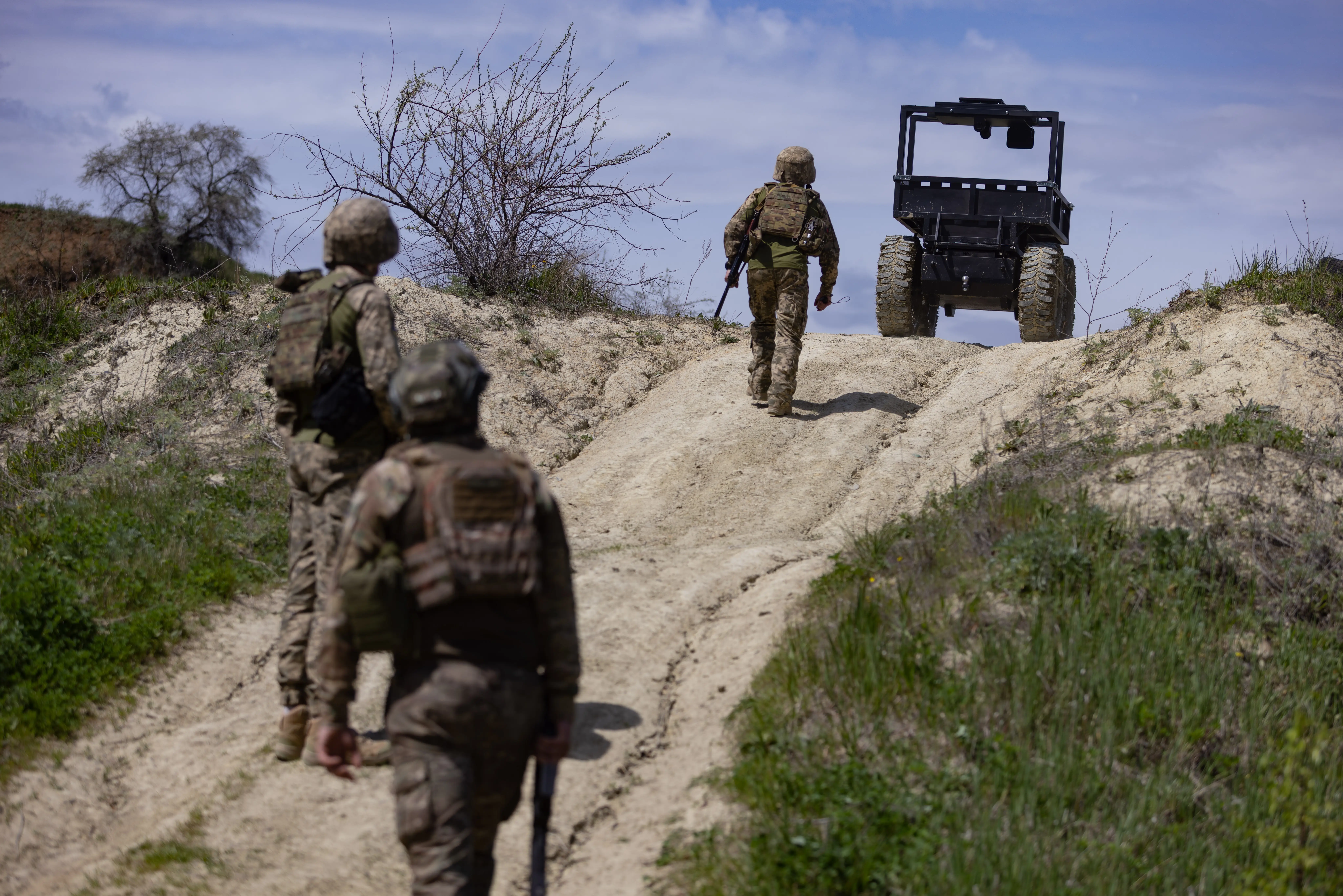 Camouflaged military personnel walk along a dirt track toward an unmanned ground vehicle on a rural hillside.