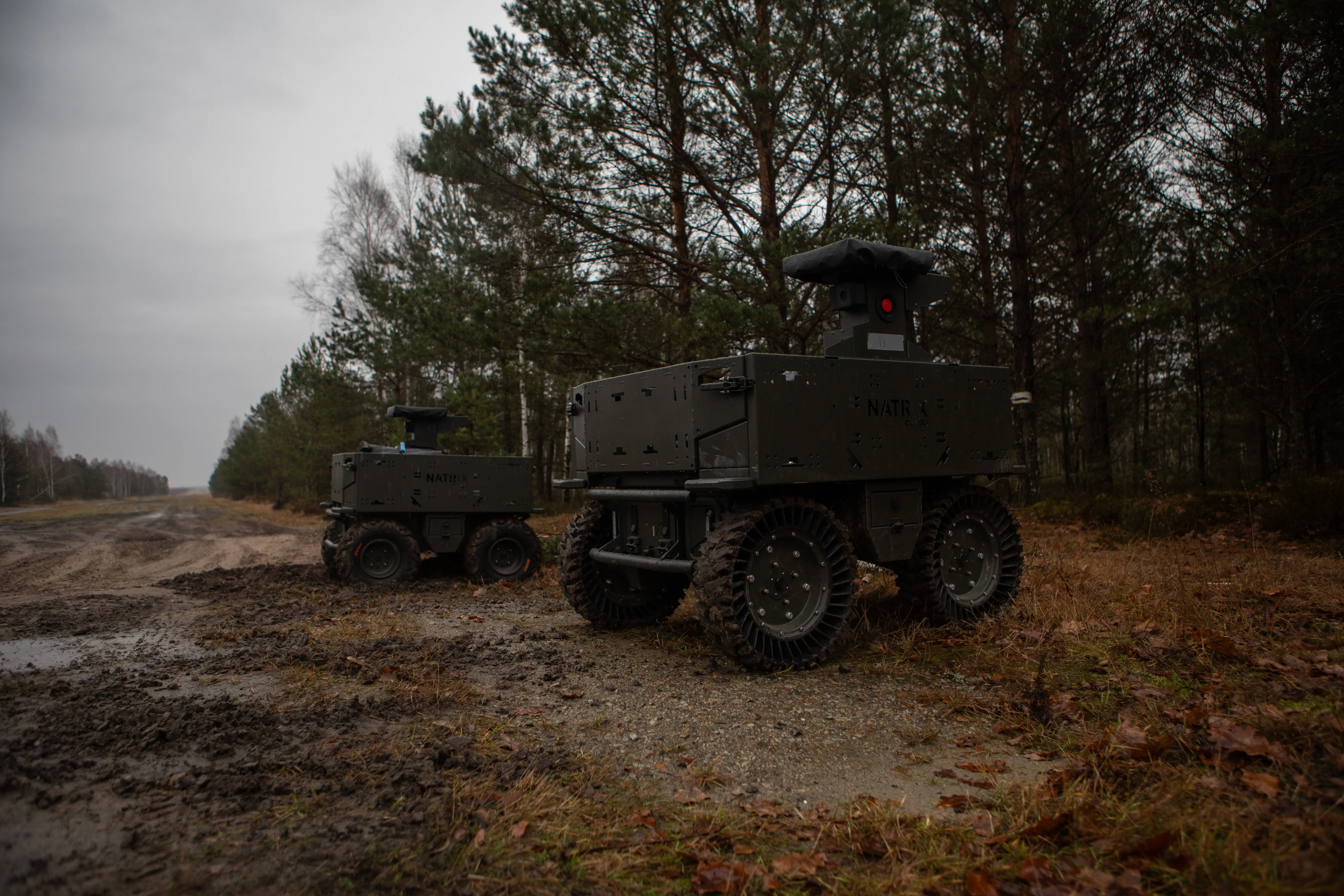Two wheeled unmanned ground vehicles sit on a muddy forest road under an overcast sky.