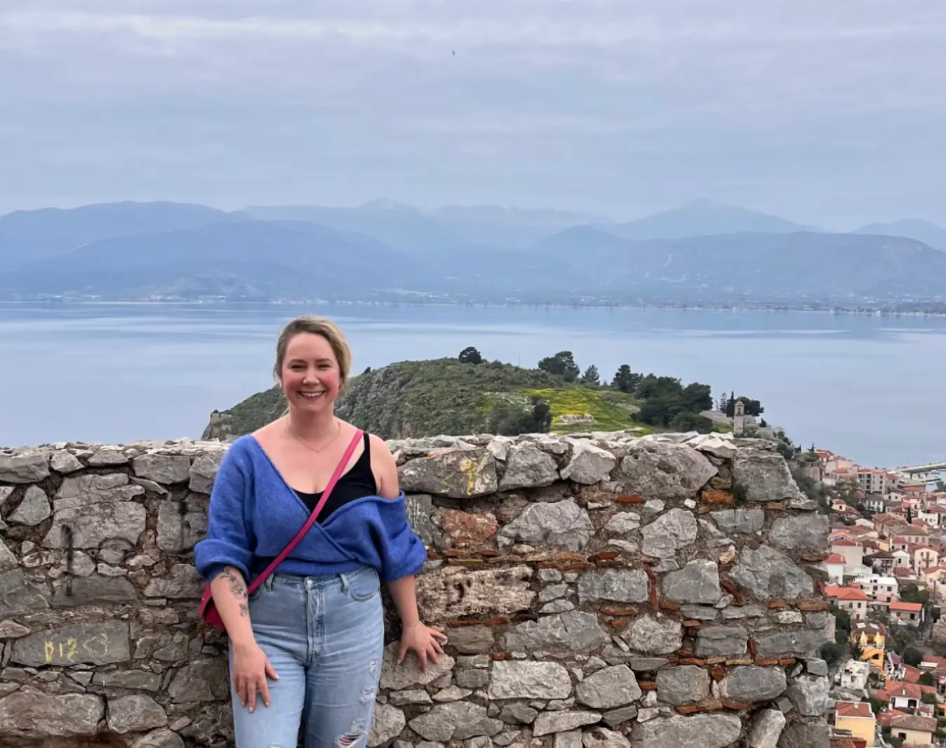 The writer wearing jeans and a sweater and standing in front of a landscape in Greece, with mountains and the water in the background.