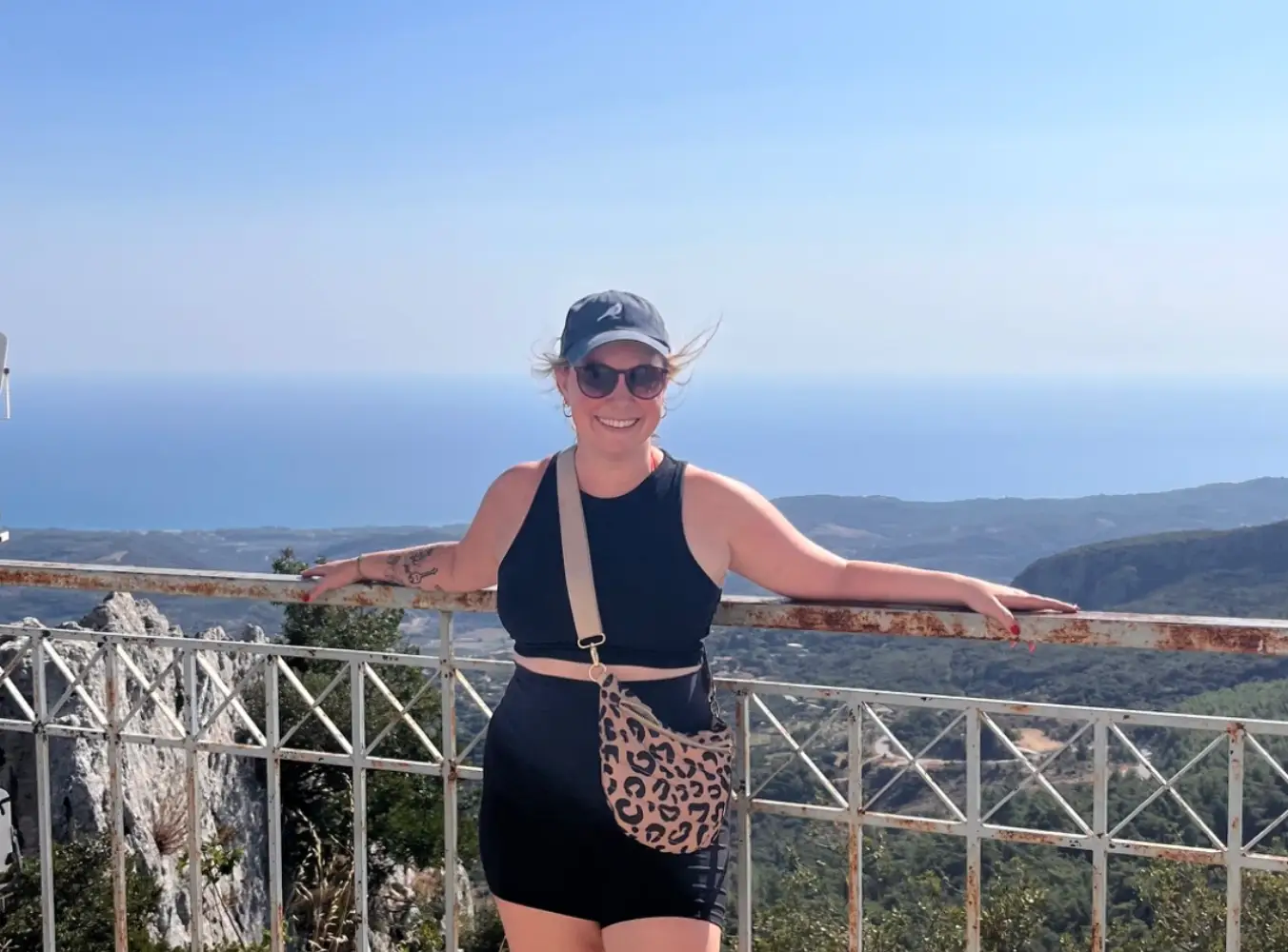The writer in sunglasses stands by a rusted railing overlooking mountains and a blue ocean coastline.