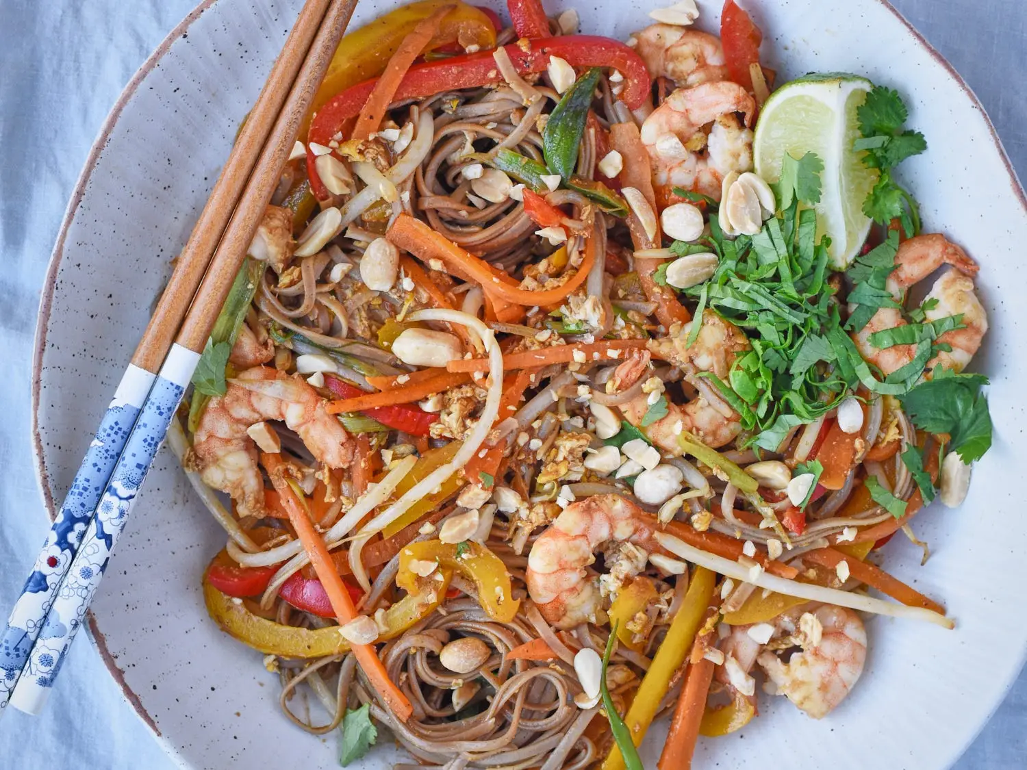 Noodle dish with shrimp, vegetables, peanuts, herbs, lime wedge, and chopsticks on a white plate.