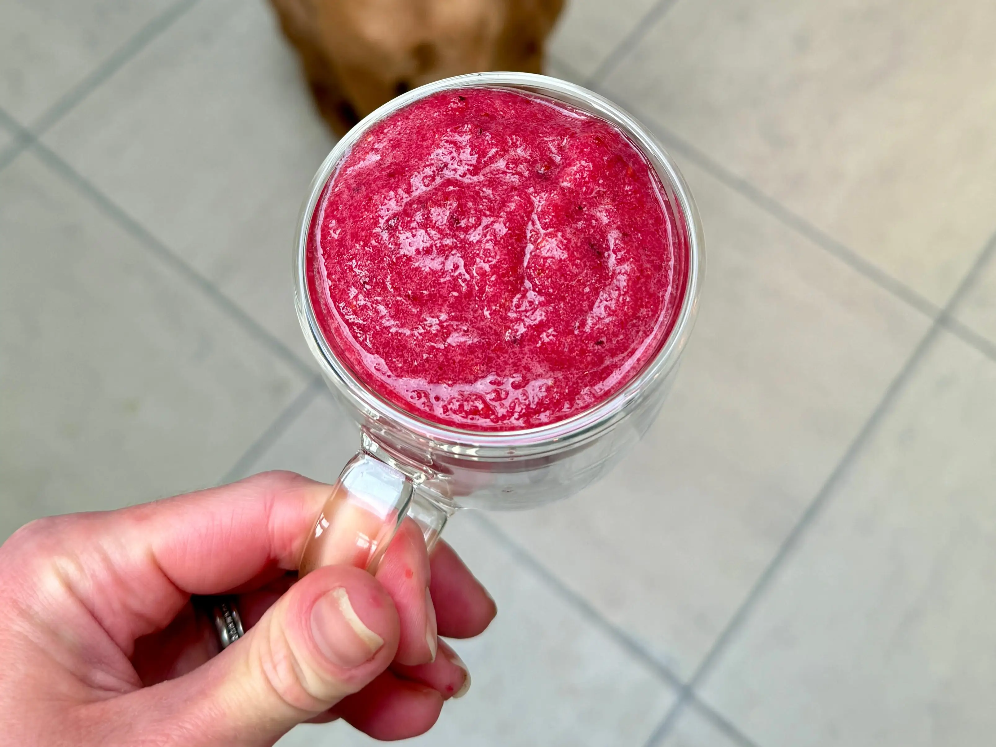 A brown dog sits on a tiled kitchen floor behind a hand holding a glass cup of bright red smoothie.
