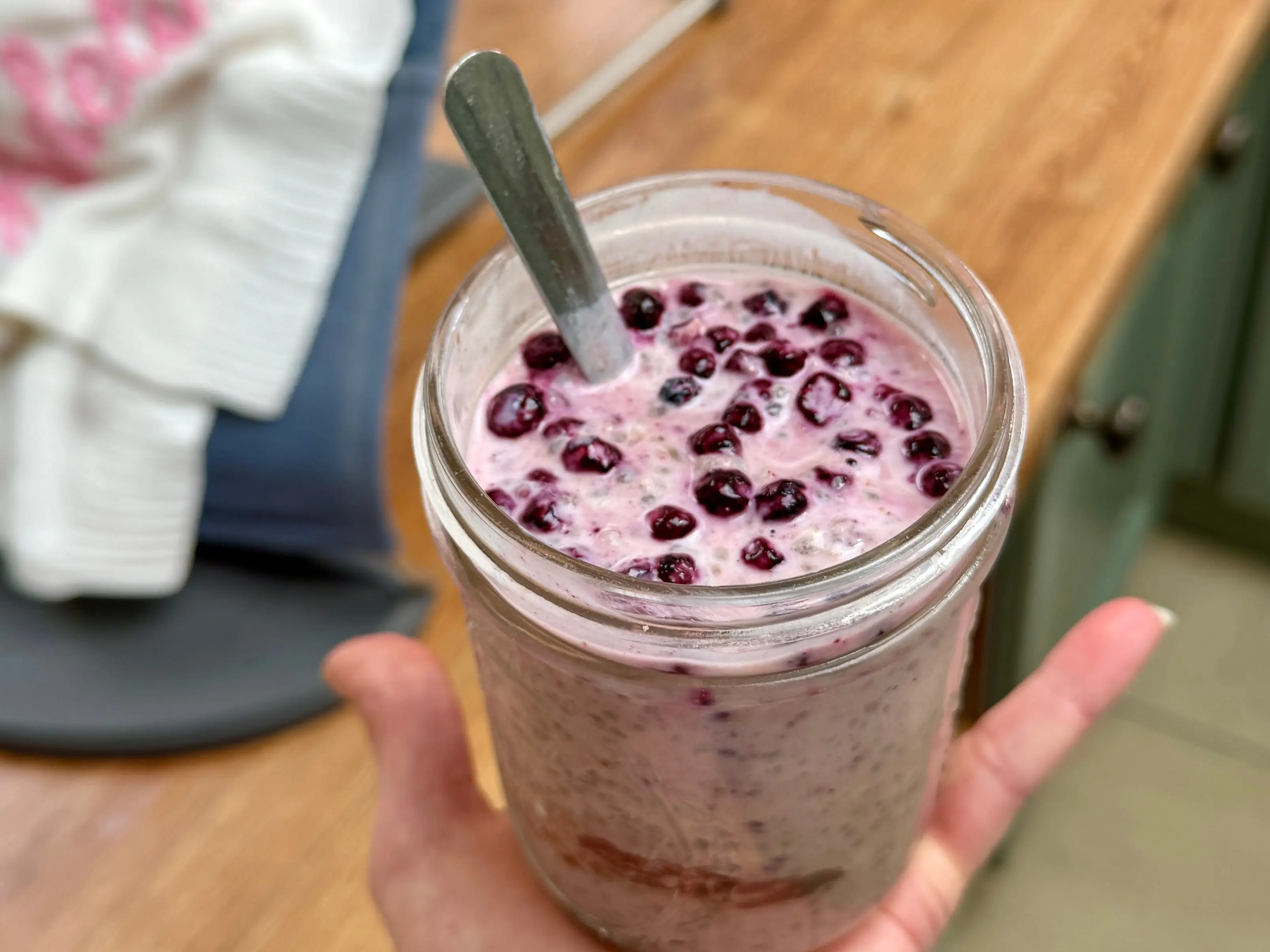 A hand holds a jar of berry-topped mixture in a kitchen with a child in a blue bouncer behind.