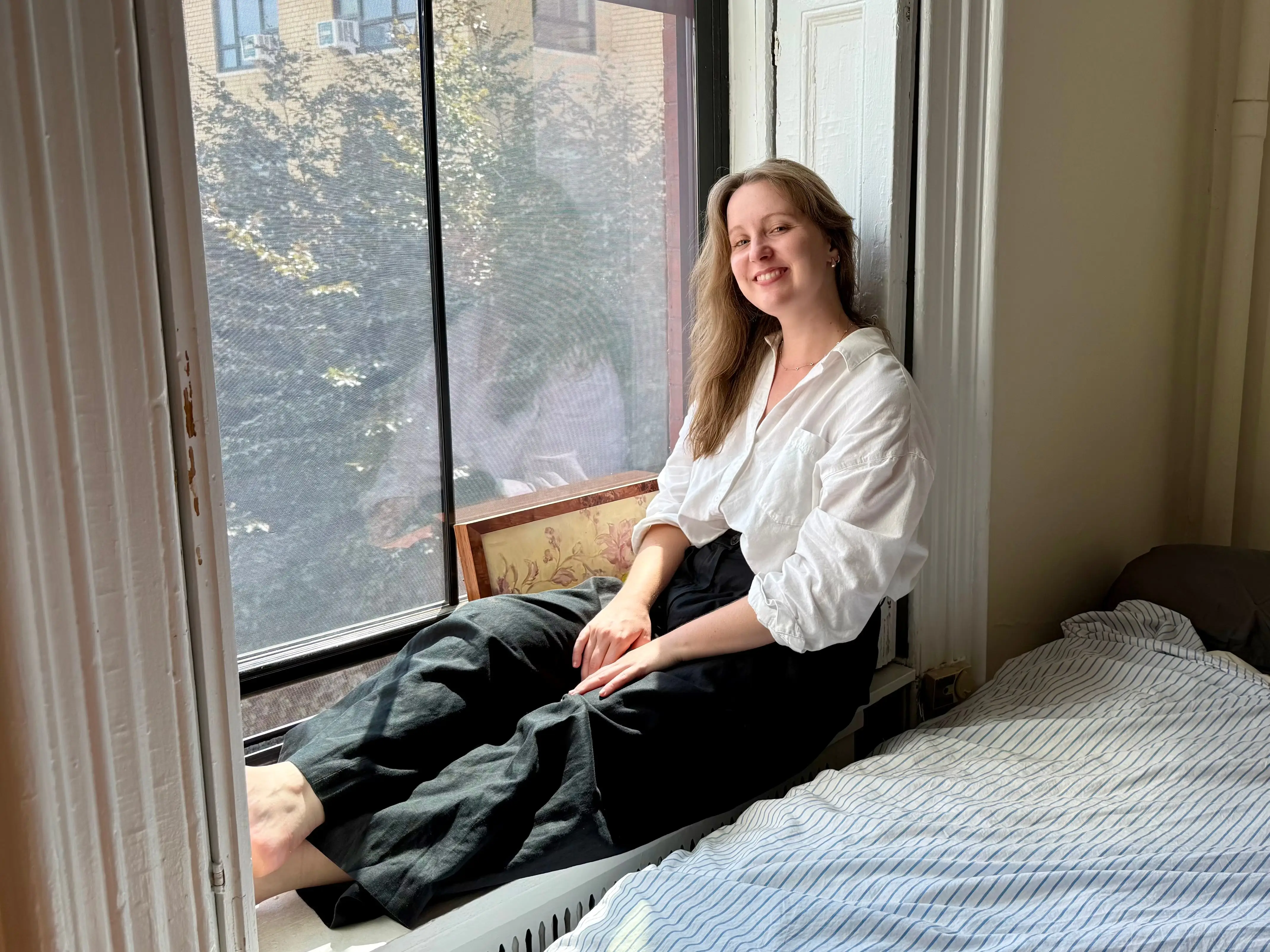 The writer sitting on the ledge of her window next to her bed in a Brooklyn apartment.