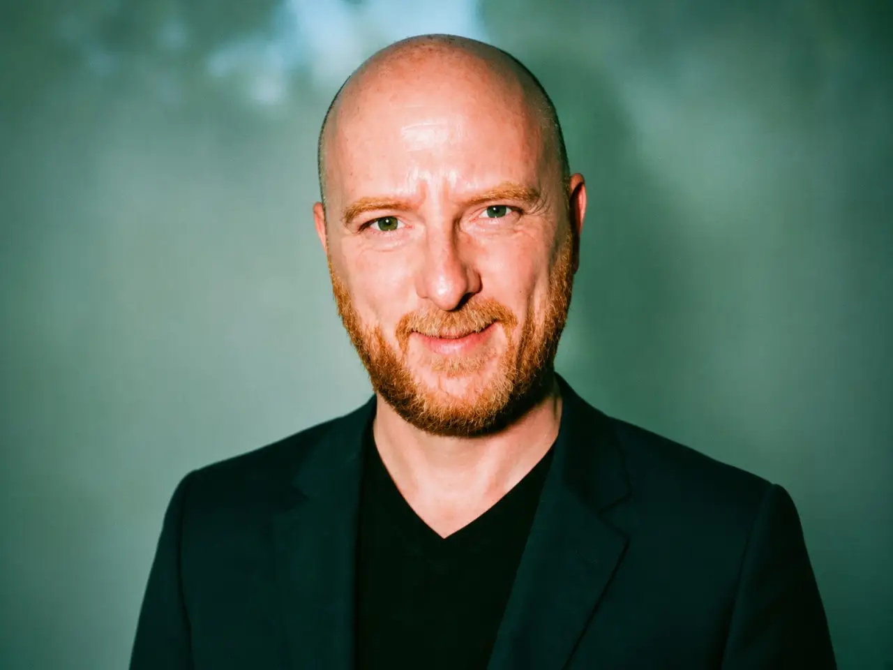 Bald person with a beard wearing a dark blazer poses against a green studio backdrop.