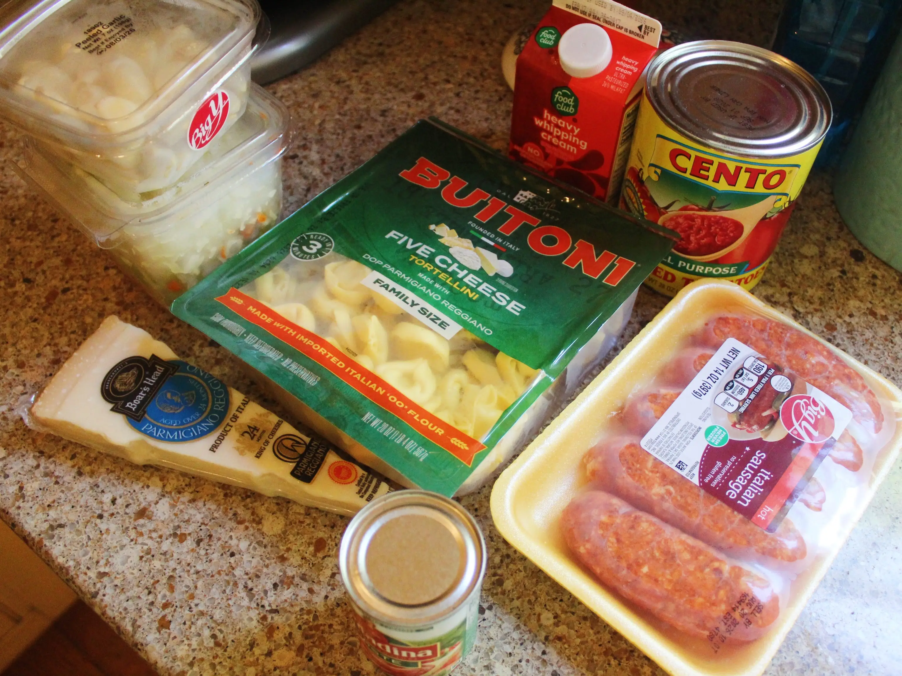 Packaged tortellini, sausage, tomatoes, cream, cheese, and chopped onions are arranged on a kitchen countertop.