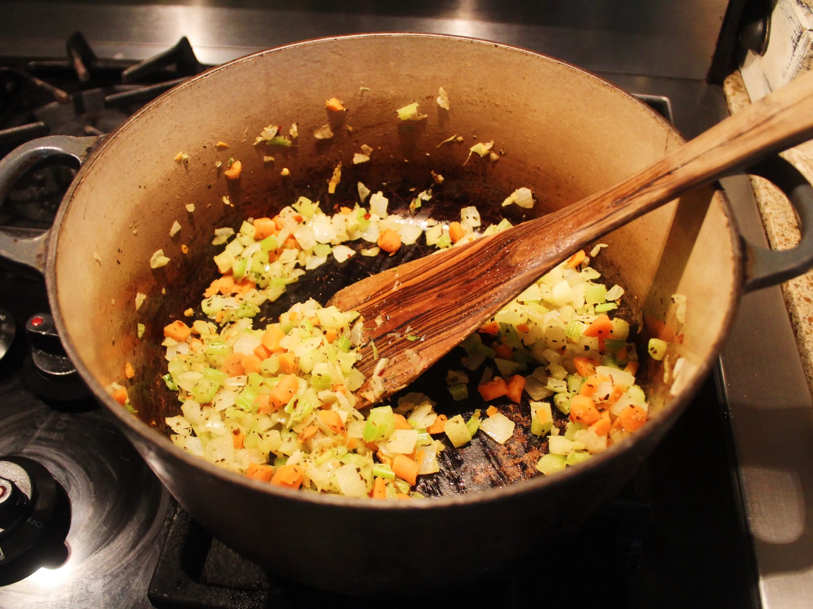 Diced vegetables sauté in a large pot on a stovetop with a wooden spoon resting inside.