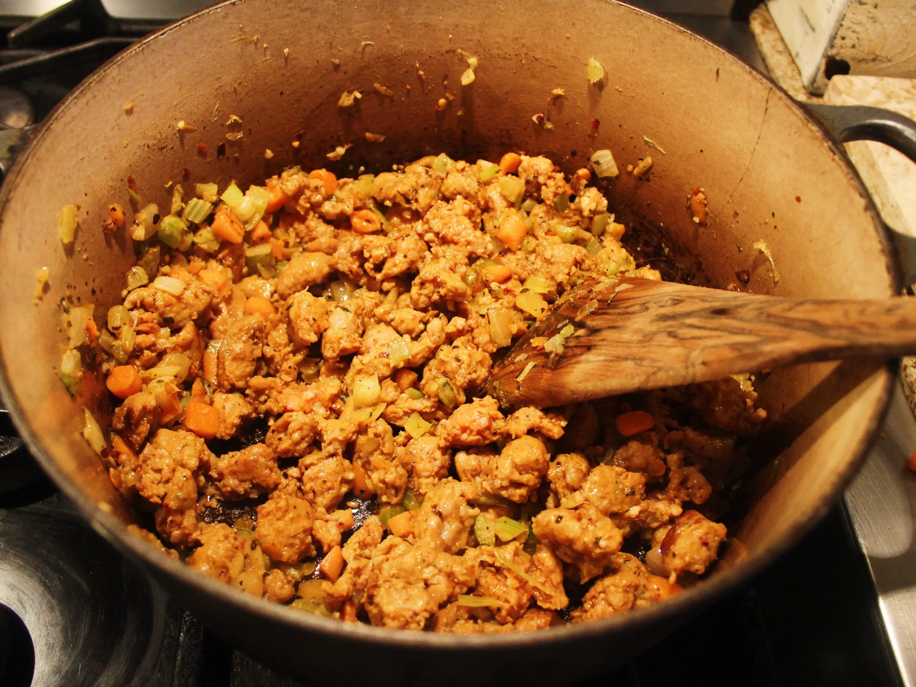 Browned ground meat and chopped vegetables cook in a large pot with a wooden spoon on a stovetop.