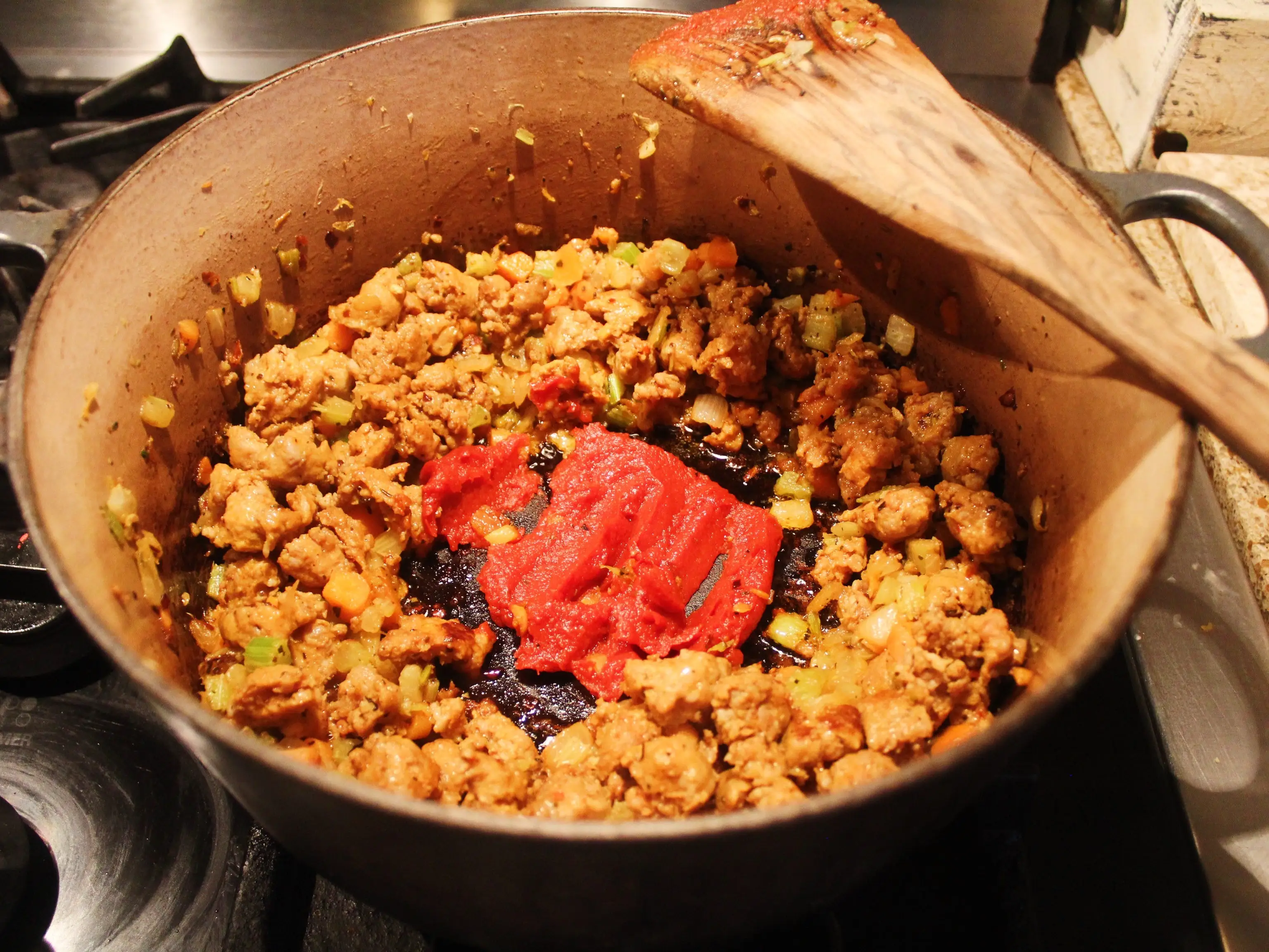 Ground meat, diced vegetables, and tomato paste cook in a large pot on a stovetop with a wooden spoon.