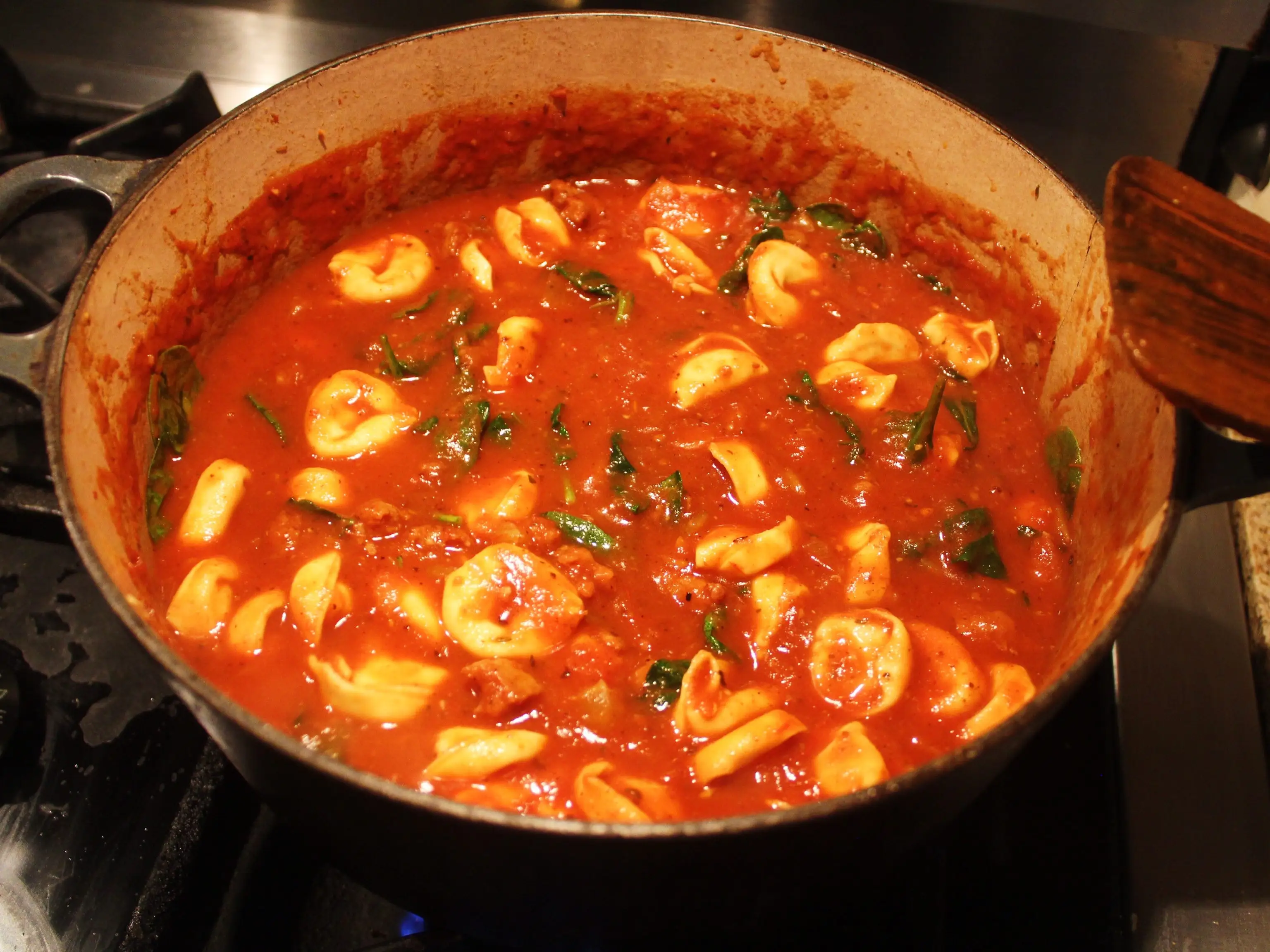 Tortellini simmer in red tomato sauce with leafy greens inside a large pot on a stovetop.