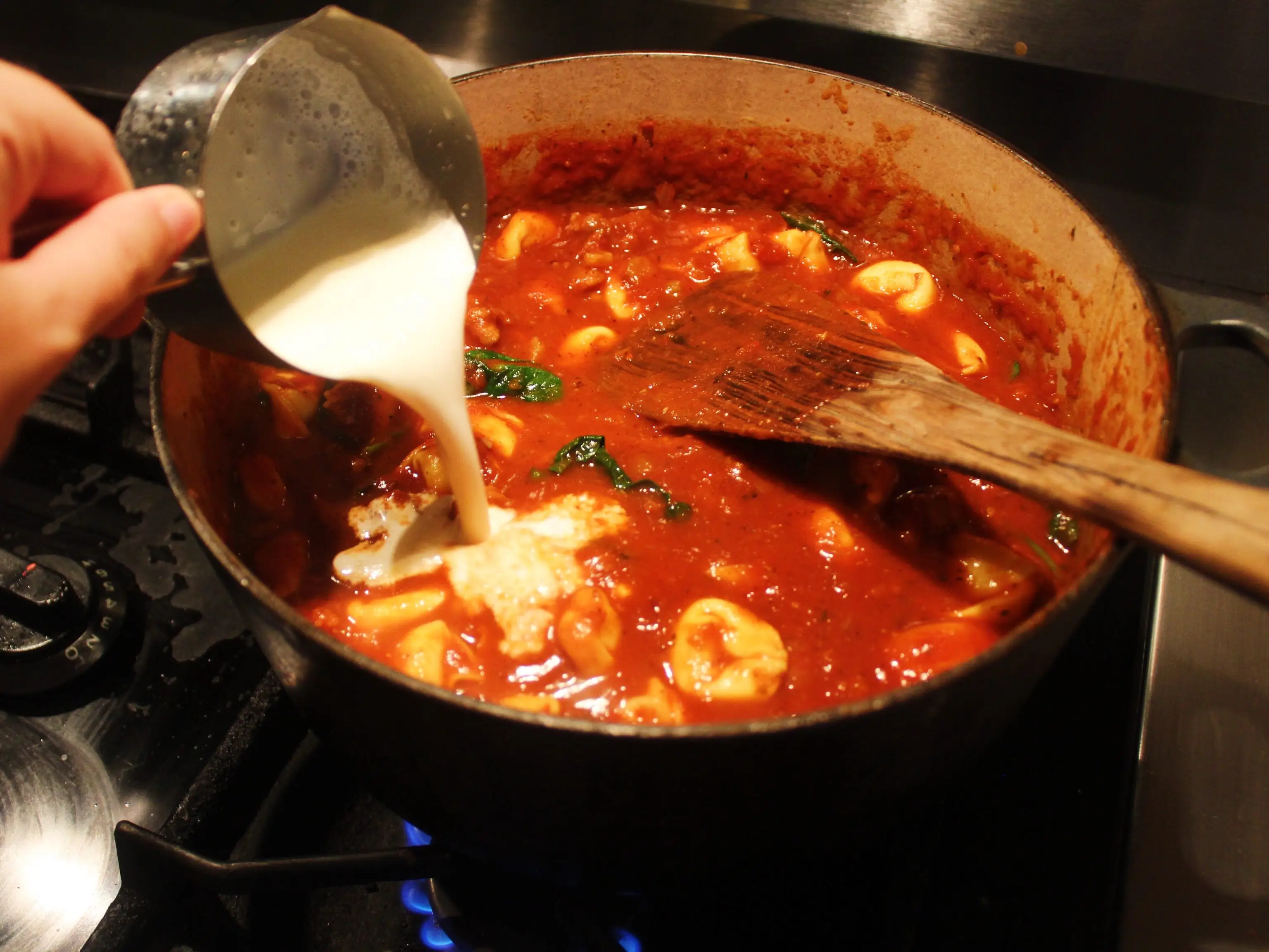 Cream is poured into a pot of red tortellini sauce simmering on a gas stovetop.