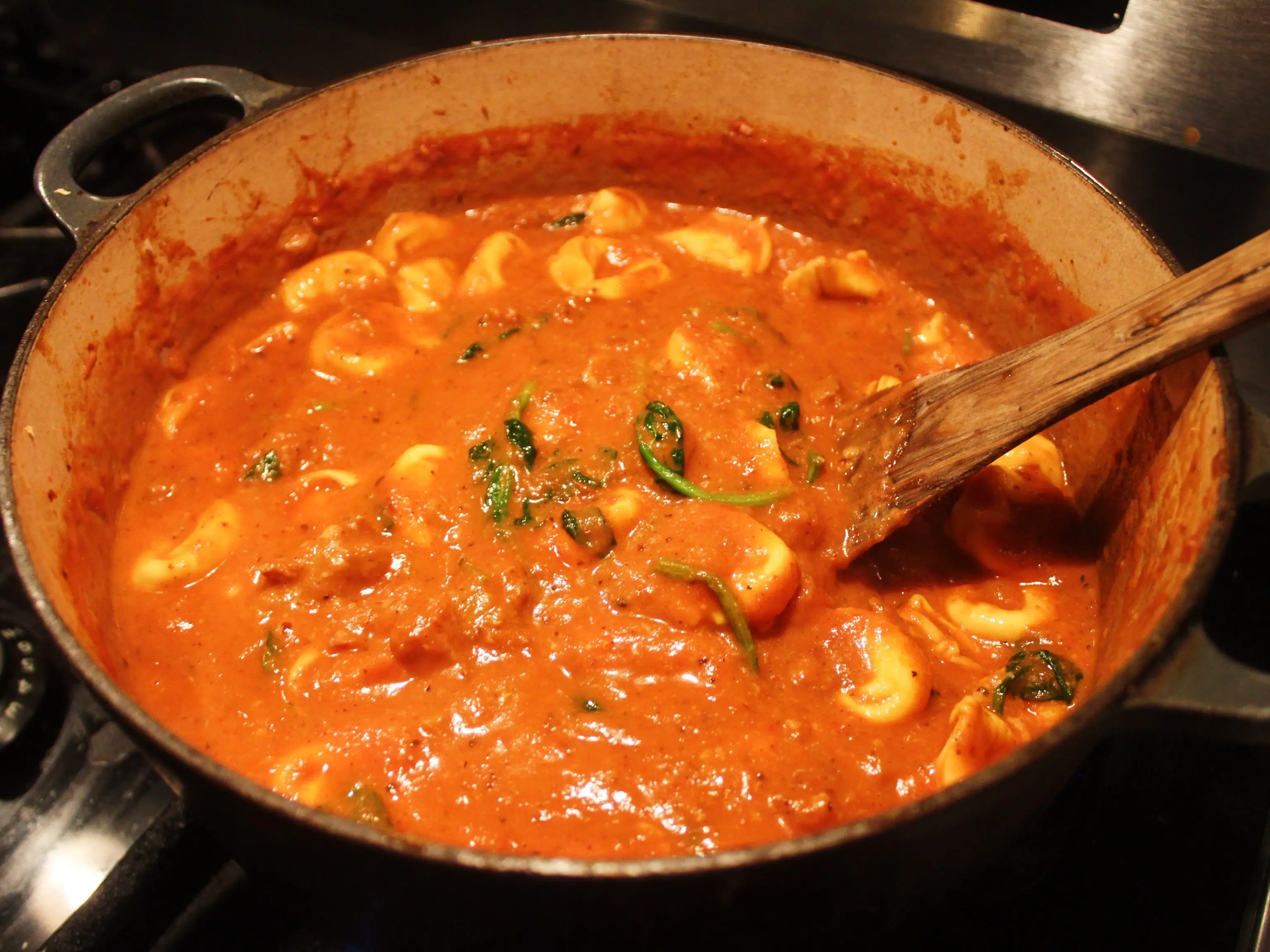Tortellini cooks in a red tomato sauce with greens in a pot on a stovetop.
