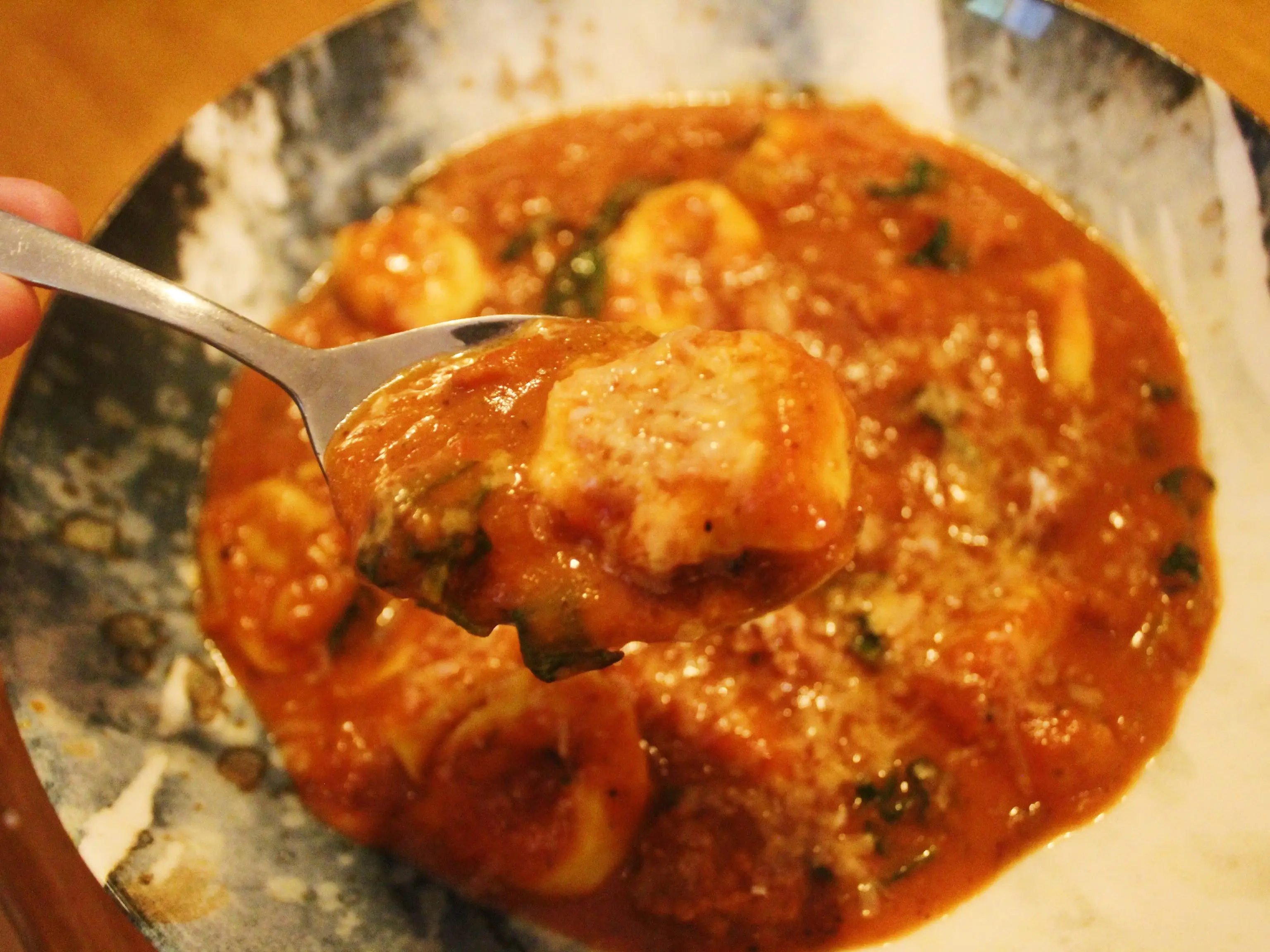 A spoon lifts a piece of pasta from a bowl of red tomato-based sauce on a wooden table.