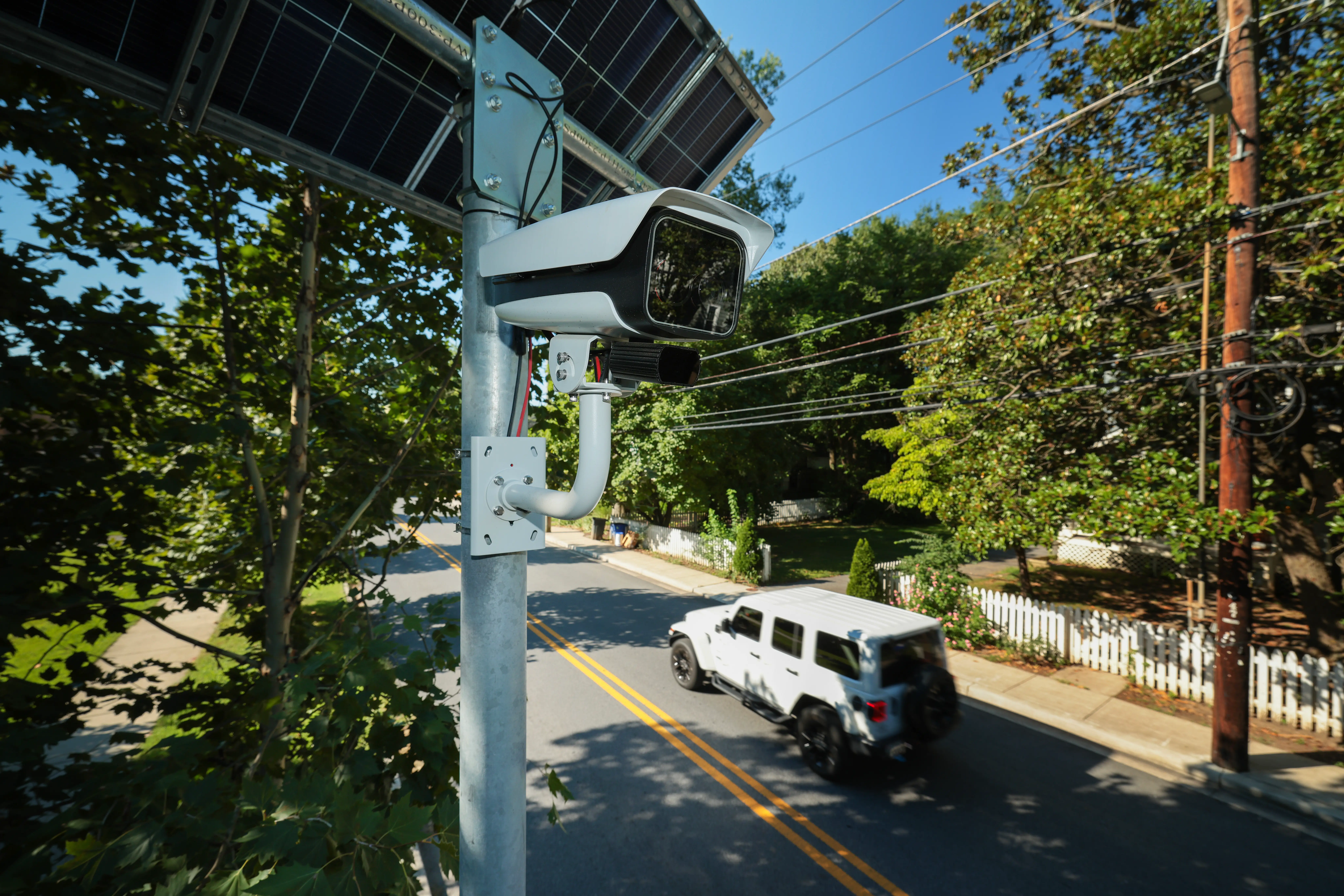 An automated license plate reader scans a road while a white Jeep Wrangler drives.