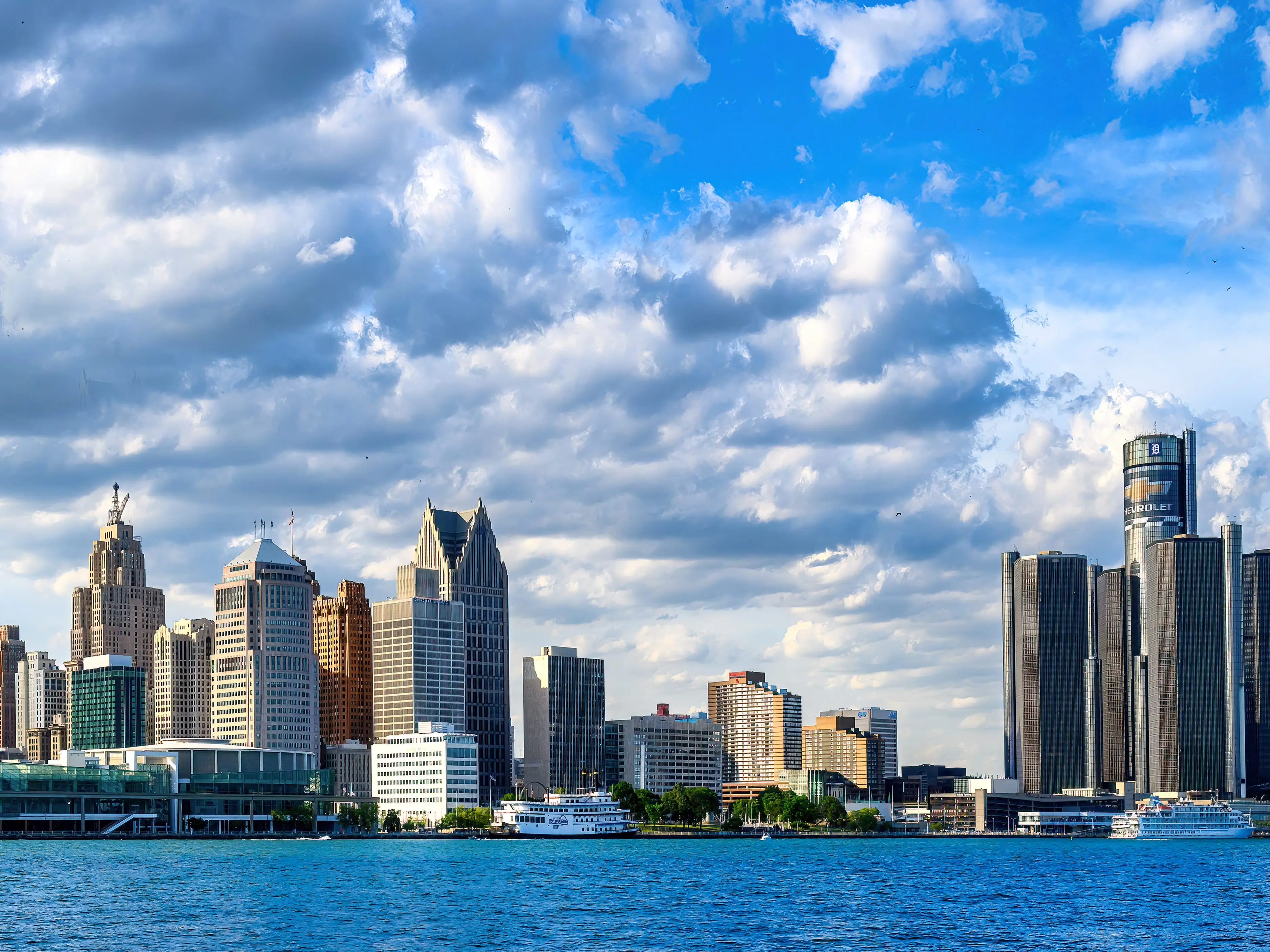 A city skyline with a river in the foreground and clouds in the background.