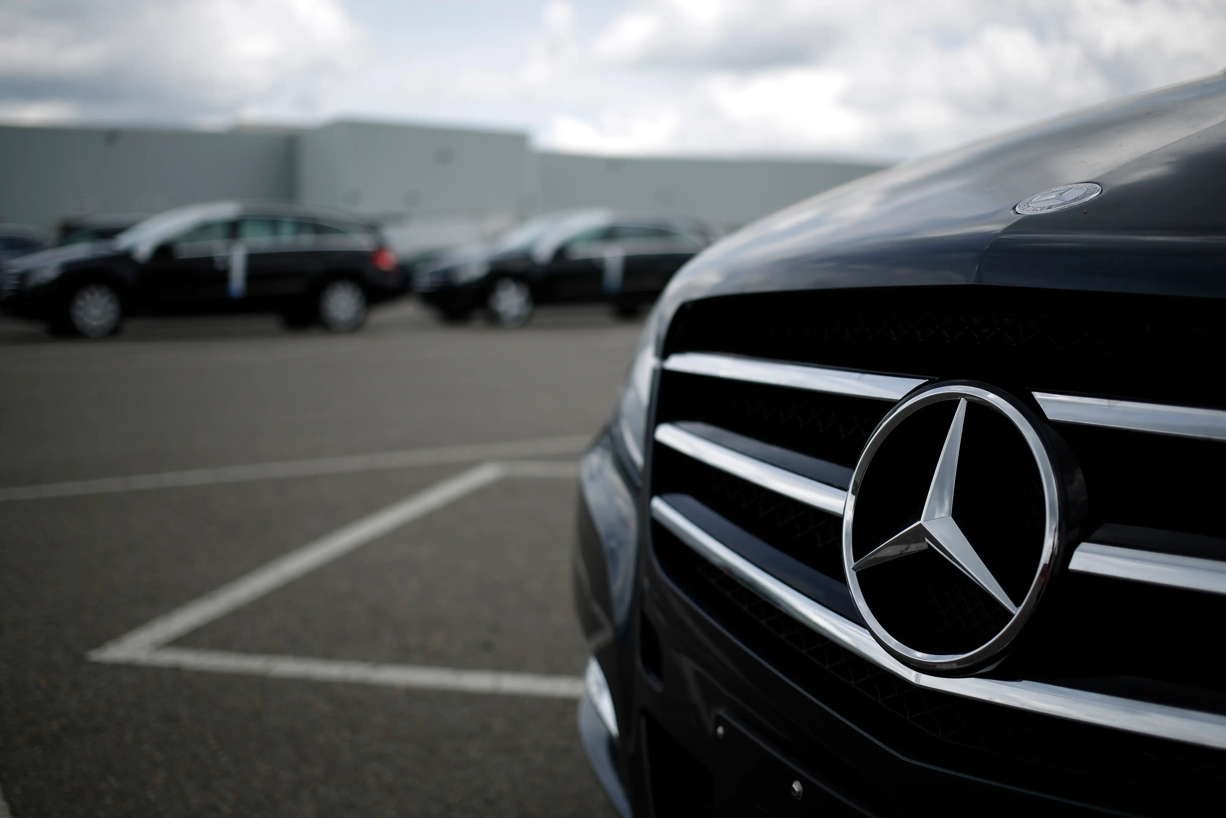 Close-up of a Mercedes-Benz grille and emblem on a black vehicle in a parking lot with other cars behind.