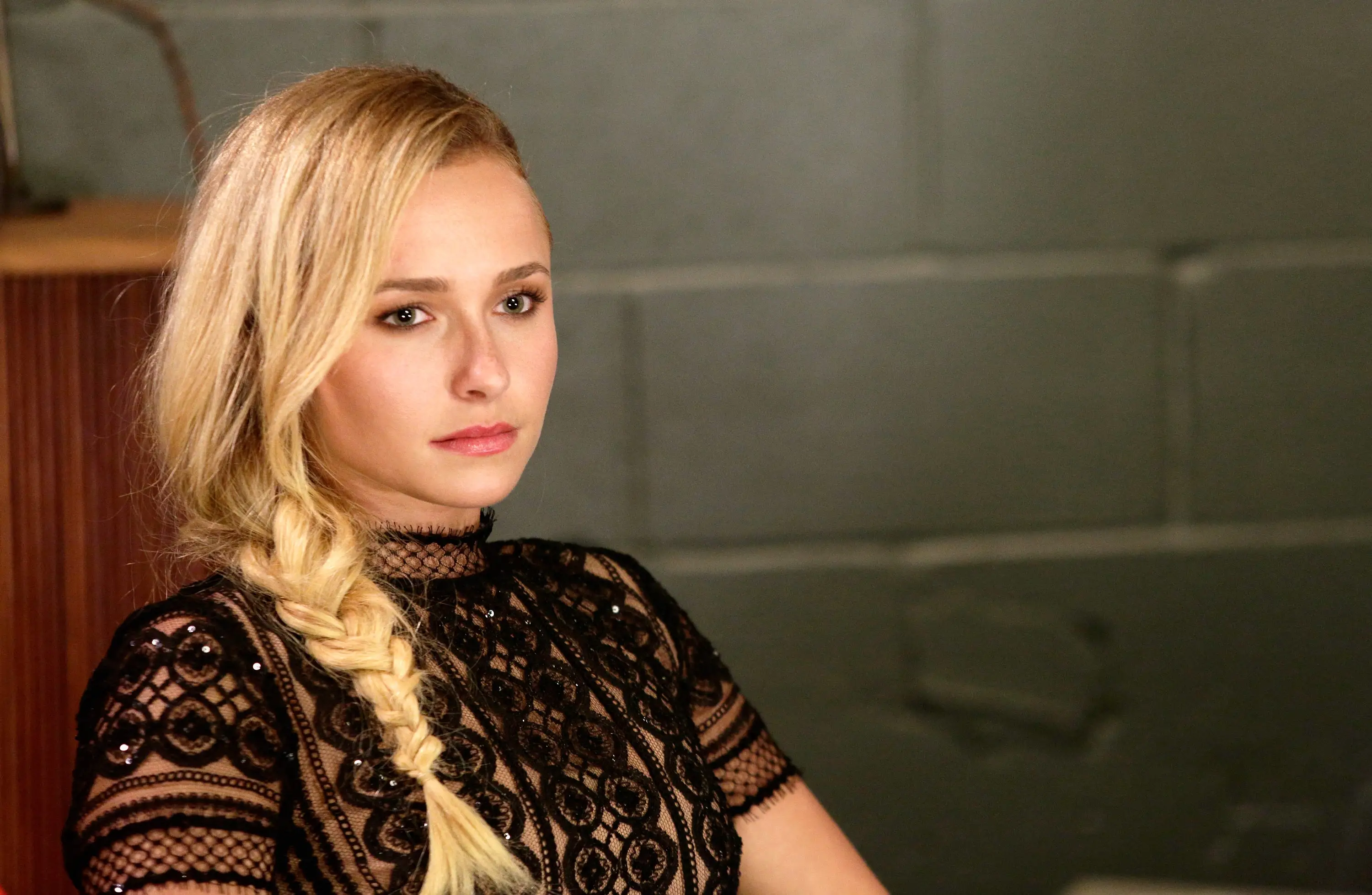 hayden panettiere with a side braid wearing a black lace top sits against a gray tiled wall.