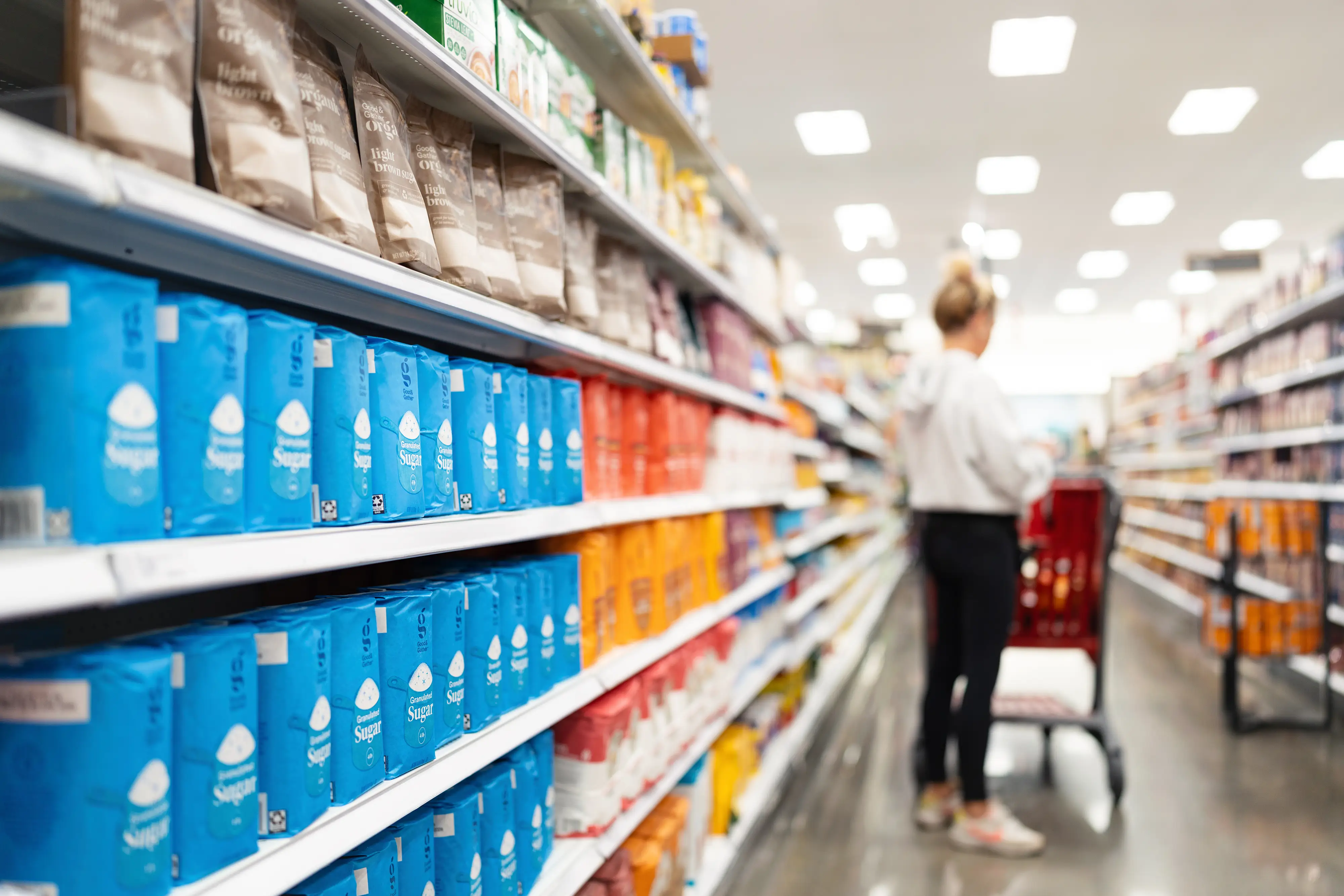 Shopper with a red cart stands in a brightly lit store aisle lined with packaged personal care products.