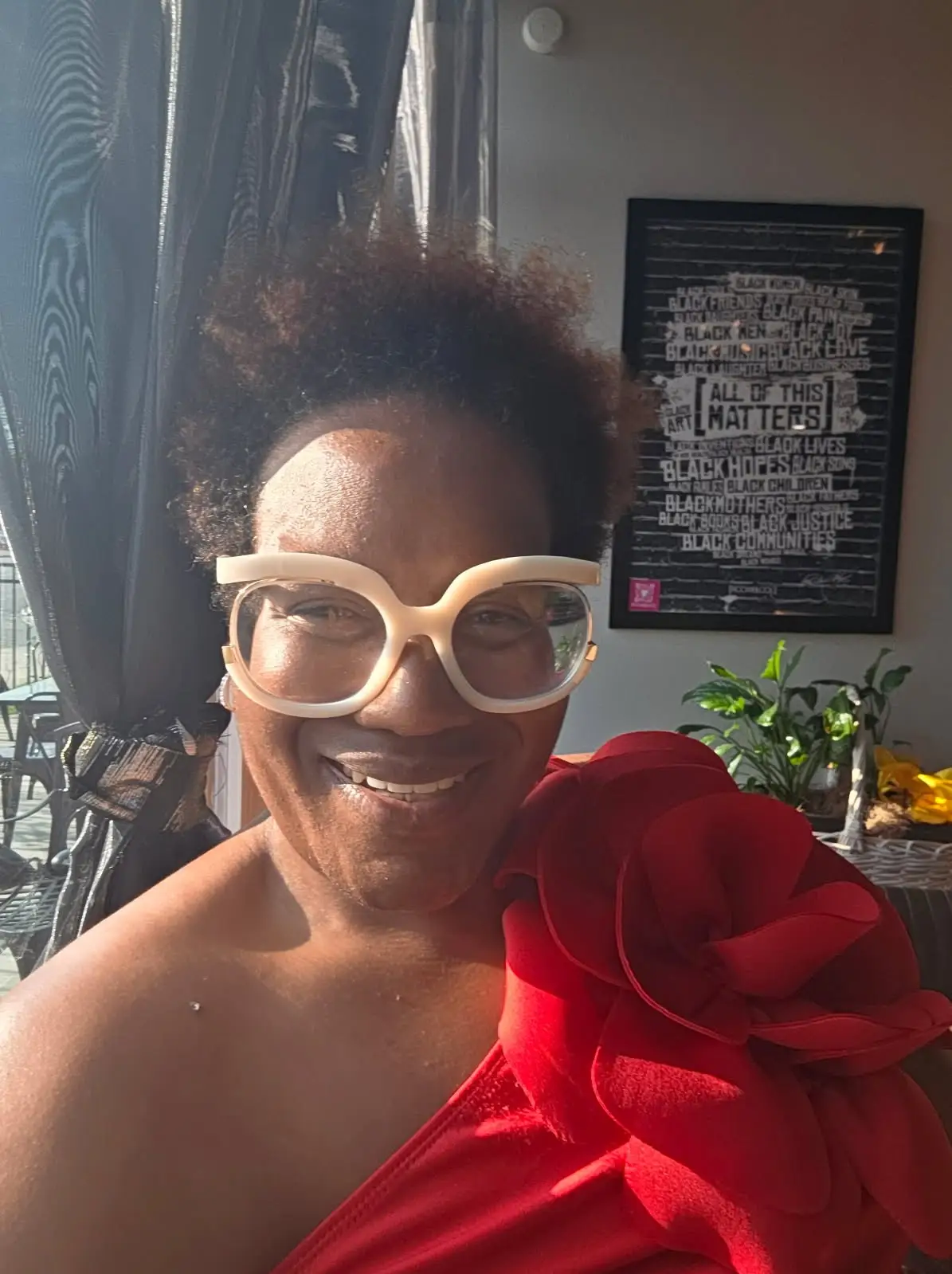 Smiling person in white glasses and a red floral-shoulder outfit stands in a sunlit room with wall art.