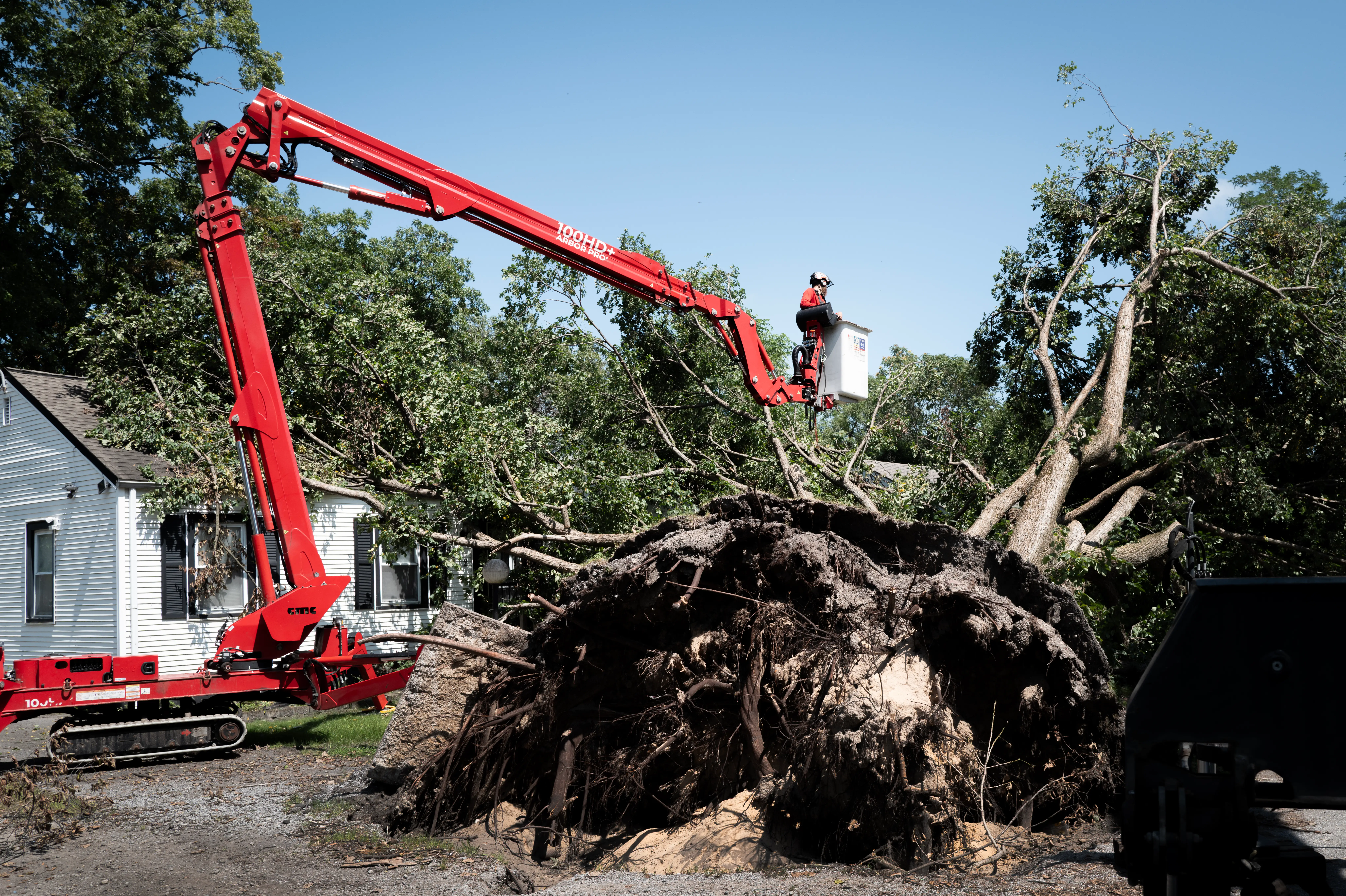 A worker in a red boom lift clears a large uprooted tree beside a house.
