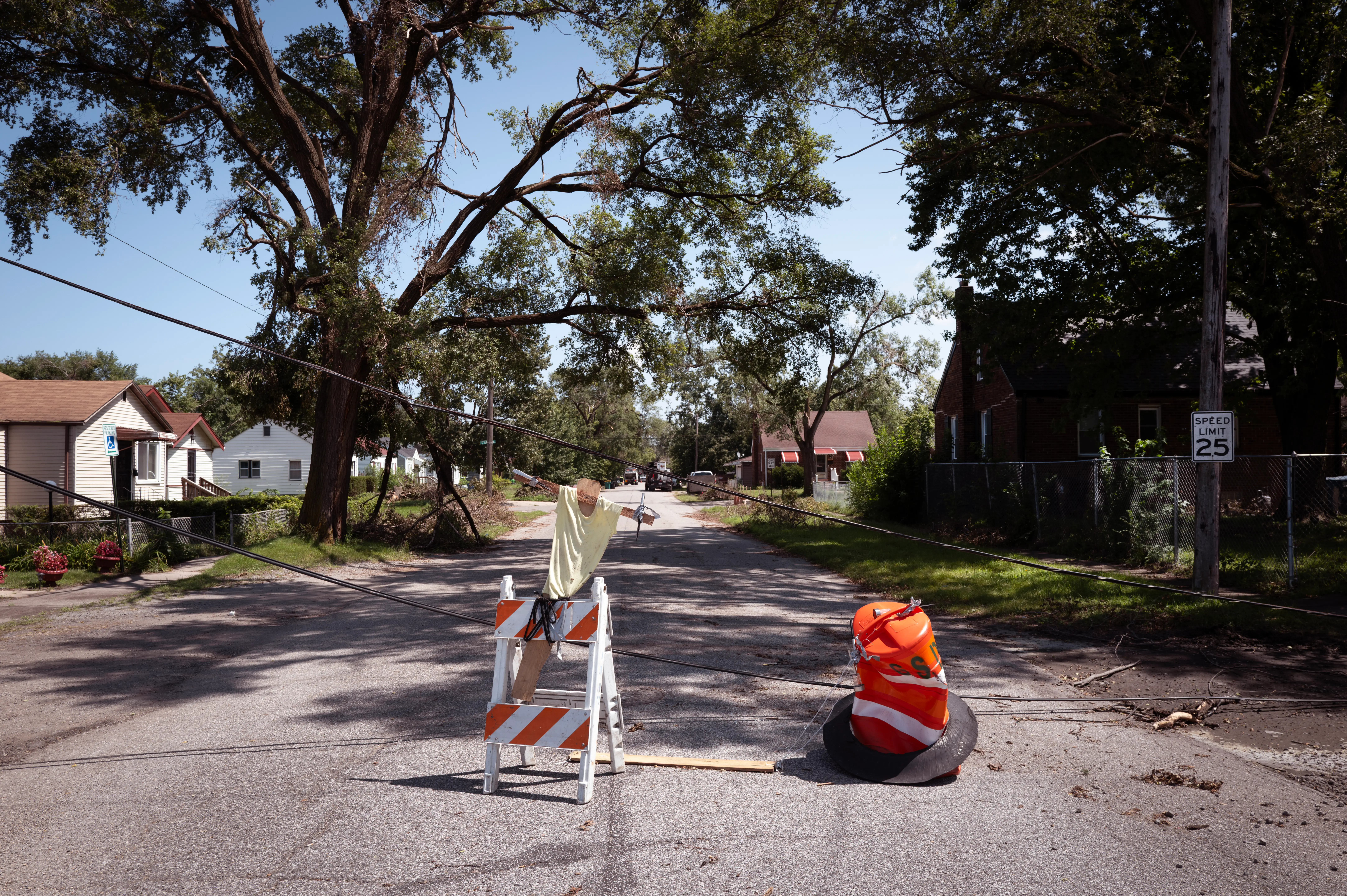 A residential street is blocked by a striped barricade and orange traffic marker near downed lines and tree debris.