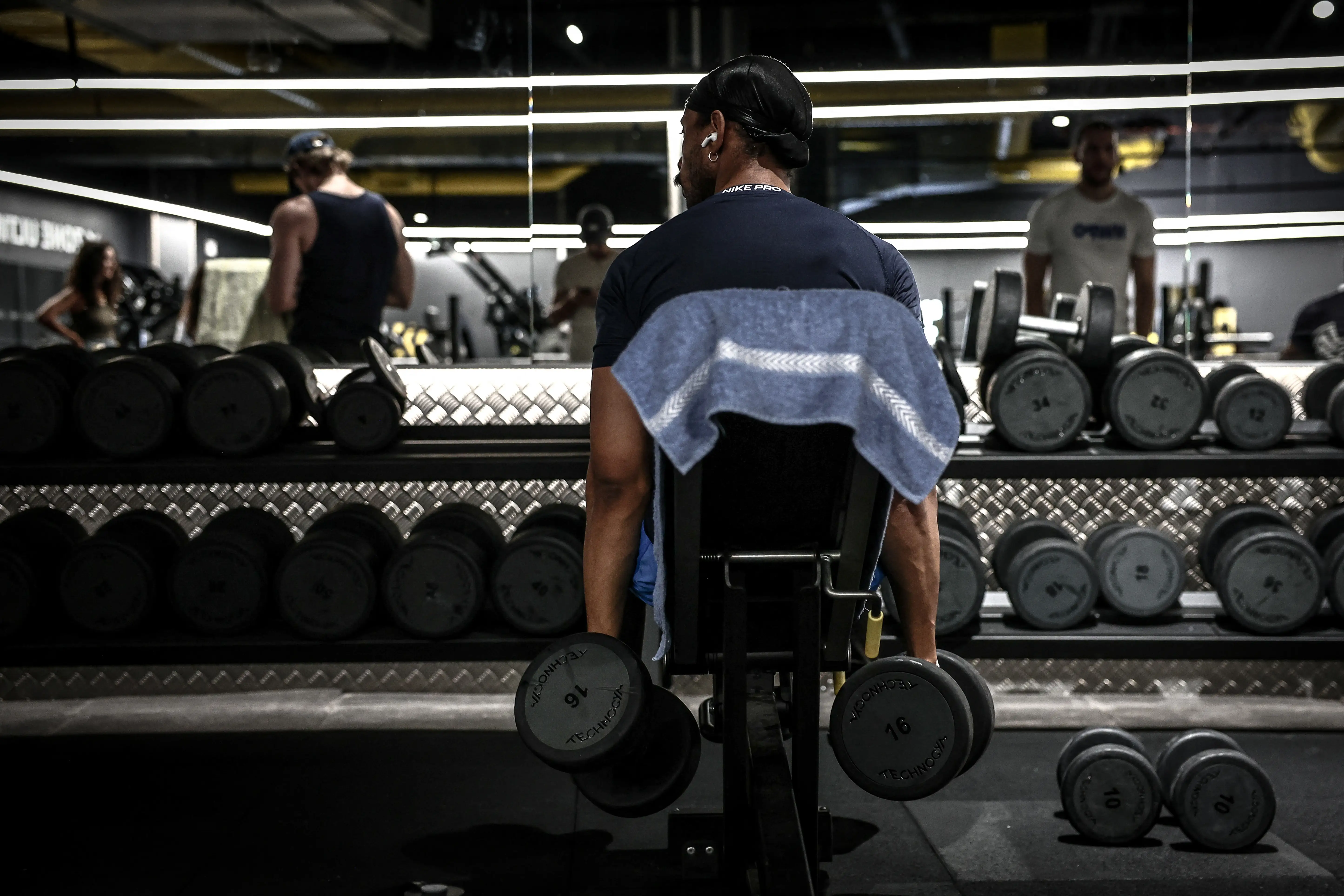Person seated at a gym bench holds dumbbells in front of a mirrored weight rack.