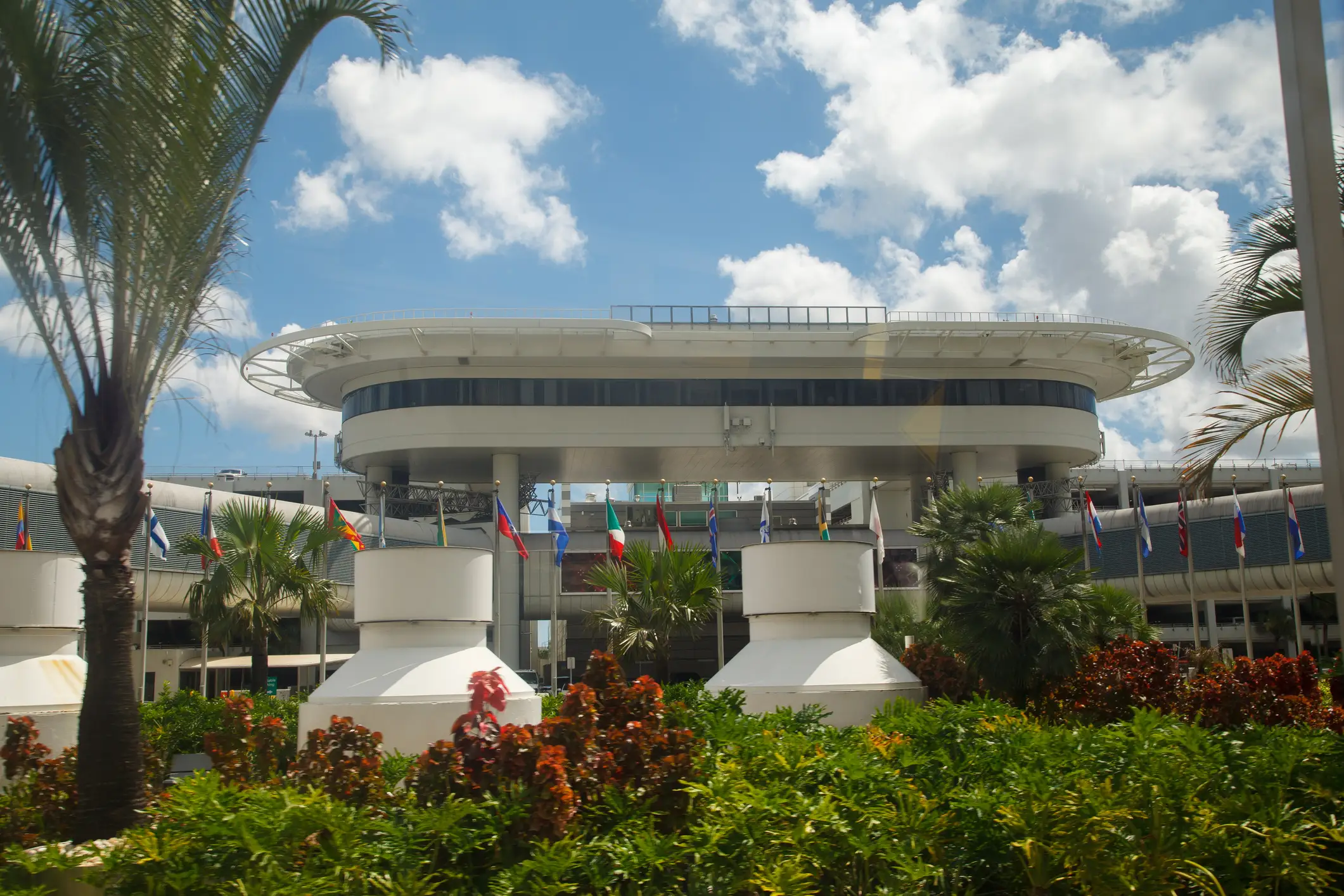 Miami International Airport with international flags, palm trees, and tropical landscaping under a partly cloudy sky.