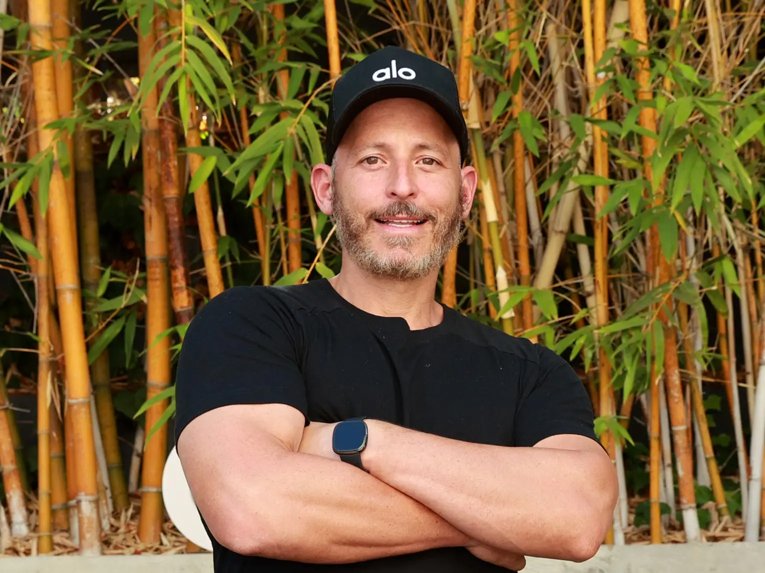 A smiling person in black shirt and blue shorts stands before bamboo and visible