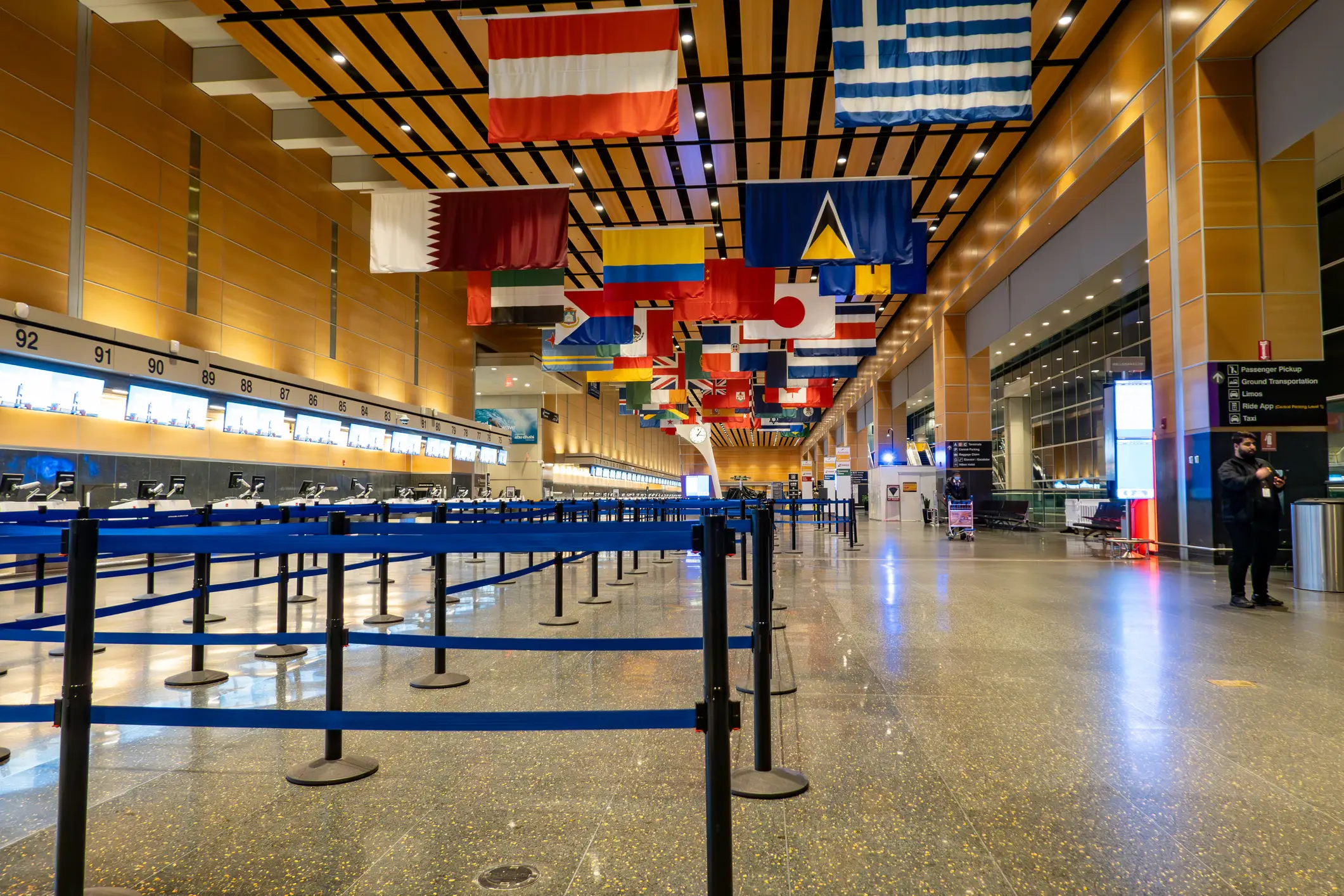 Airport check-in hall with empty queue barriers, numbered counters, and international flags hanging overhead.