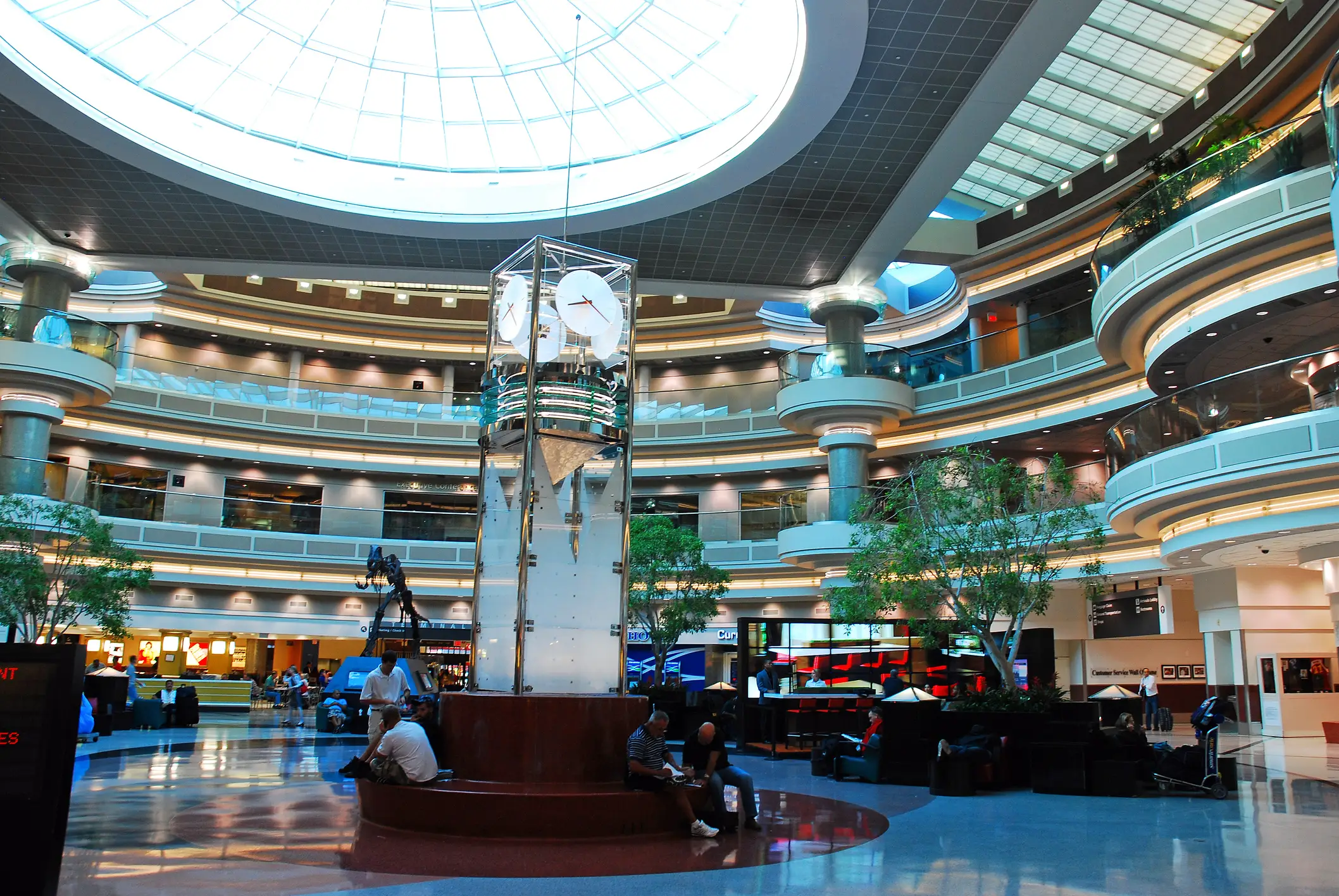 Multi-level indoor atrium at Hartsfield-Jackson Atlanta International Airport with a skylight, central clock tower, seating areas, trees, and travelers with luggage.
