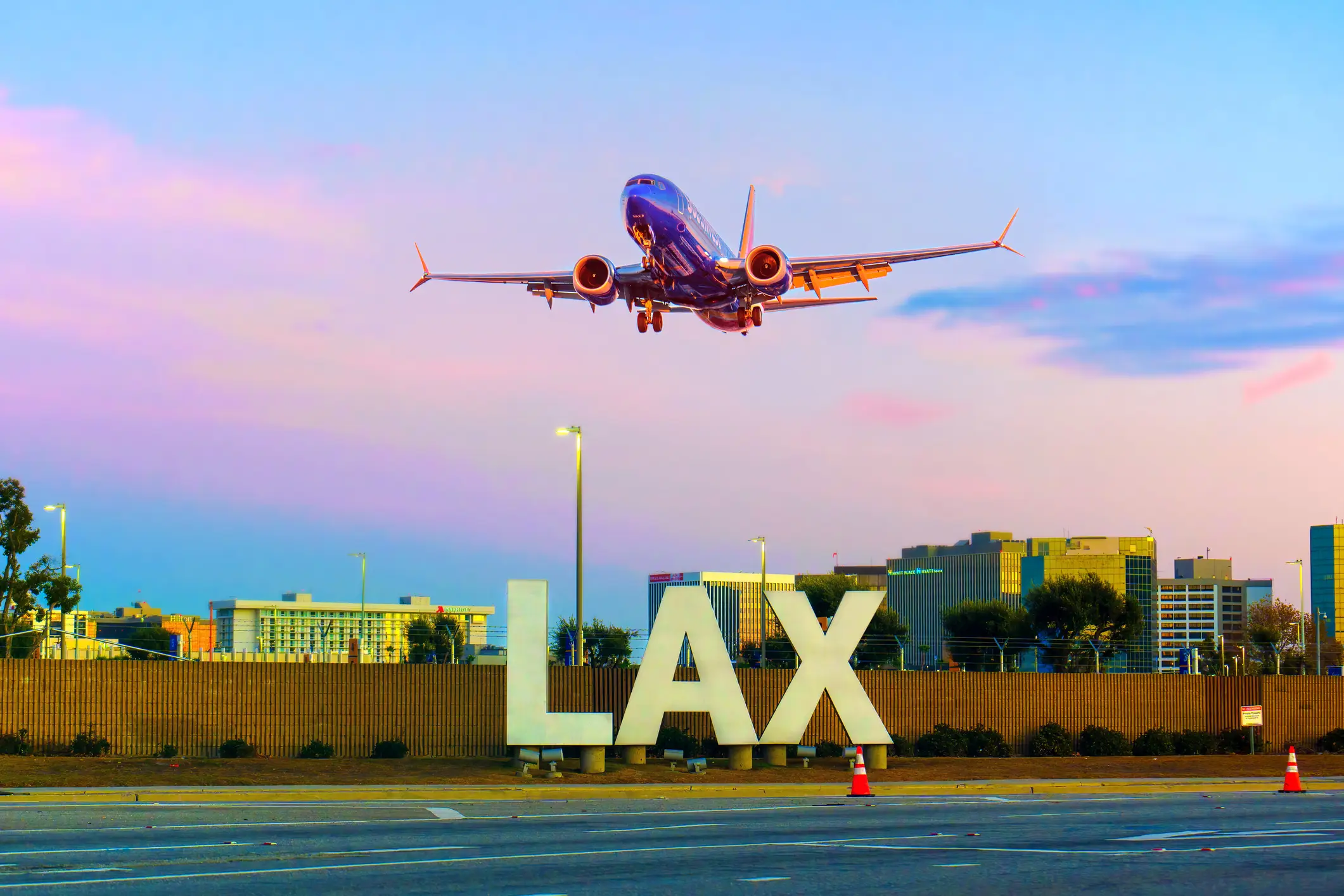 Commercial jet descends over a large LAX sign near airport roadways at colorful sunset.