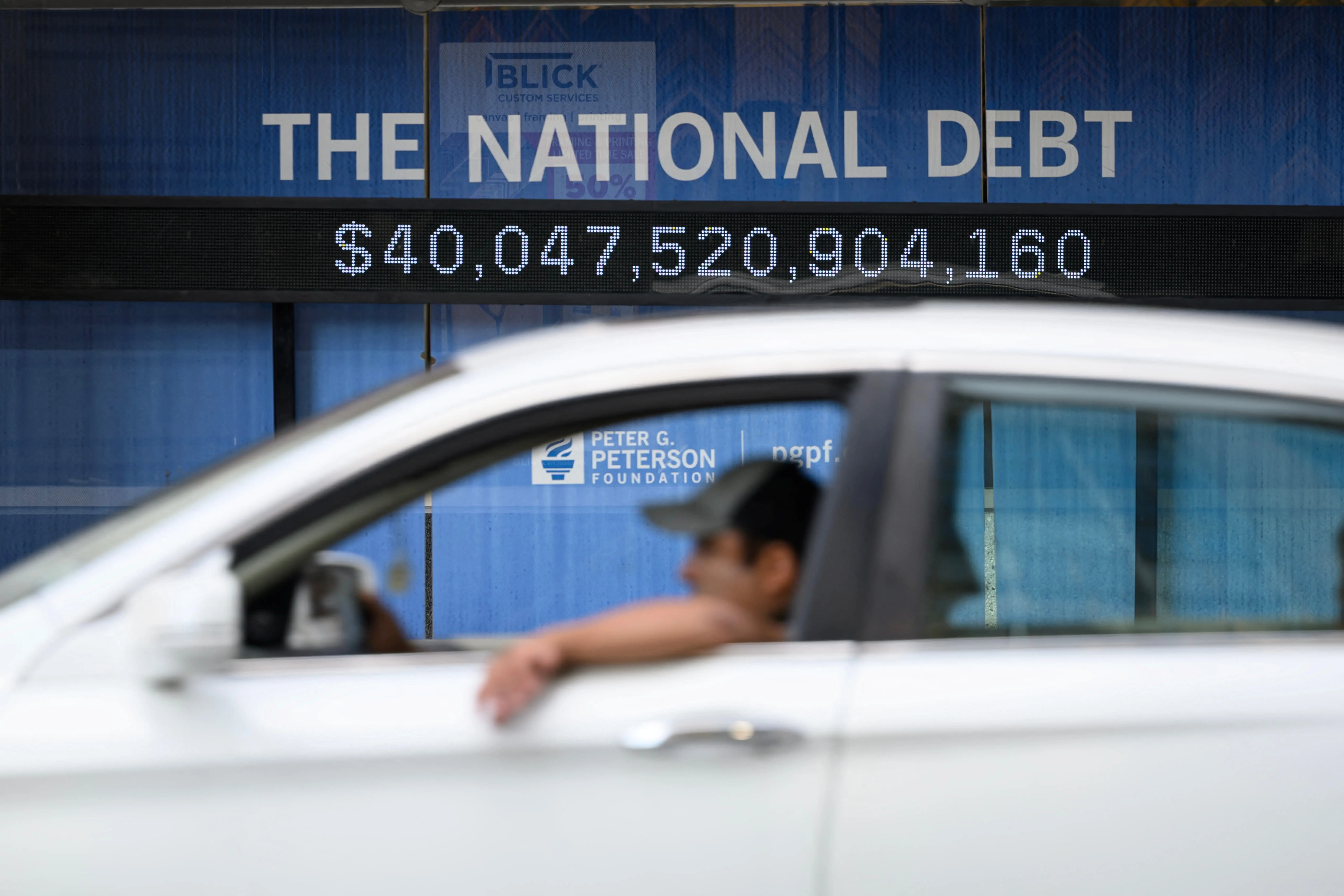 A white car passes a digital “THE NATIONAL DEBT” sign showing $40,047,520,904,160.