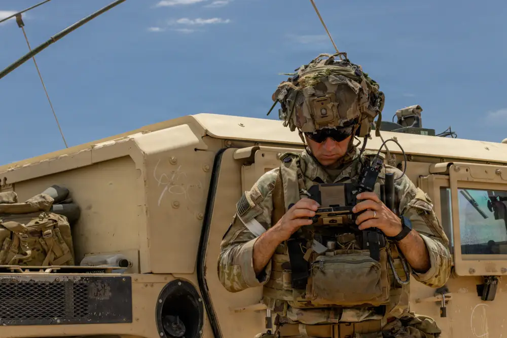 A US Army soldier looks down at their End User Device. A truck is parked in the background.