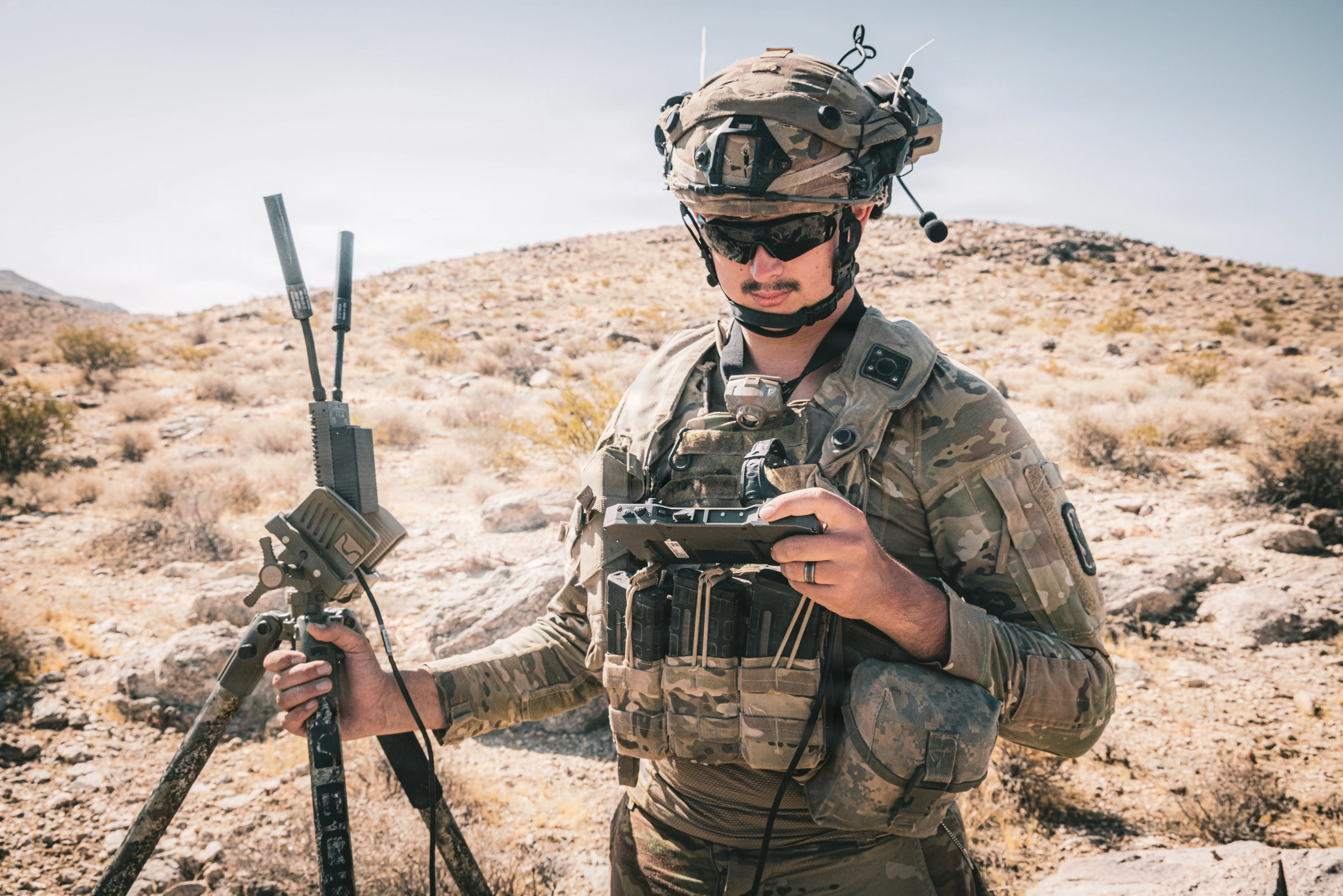 A US Army soldier looks down at their End User Device while holding an antenna for a drone in the desert.